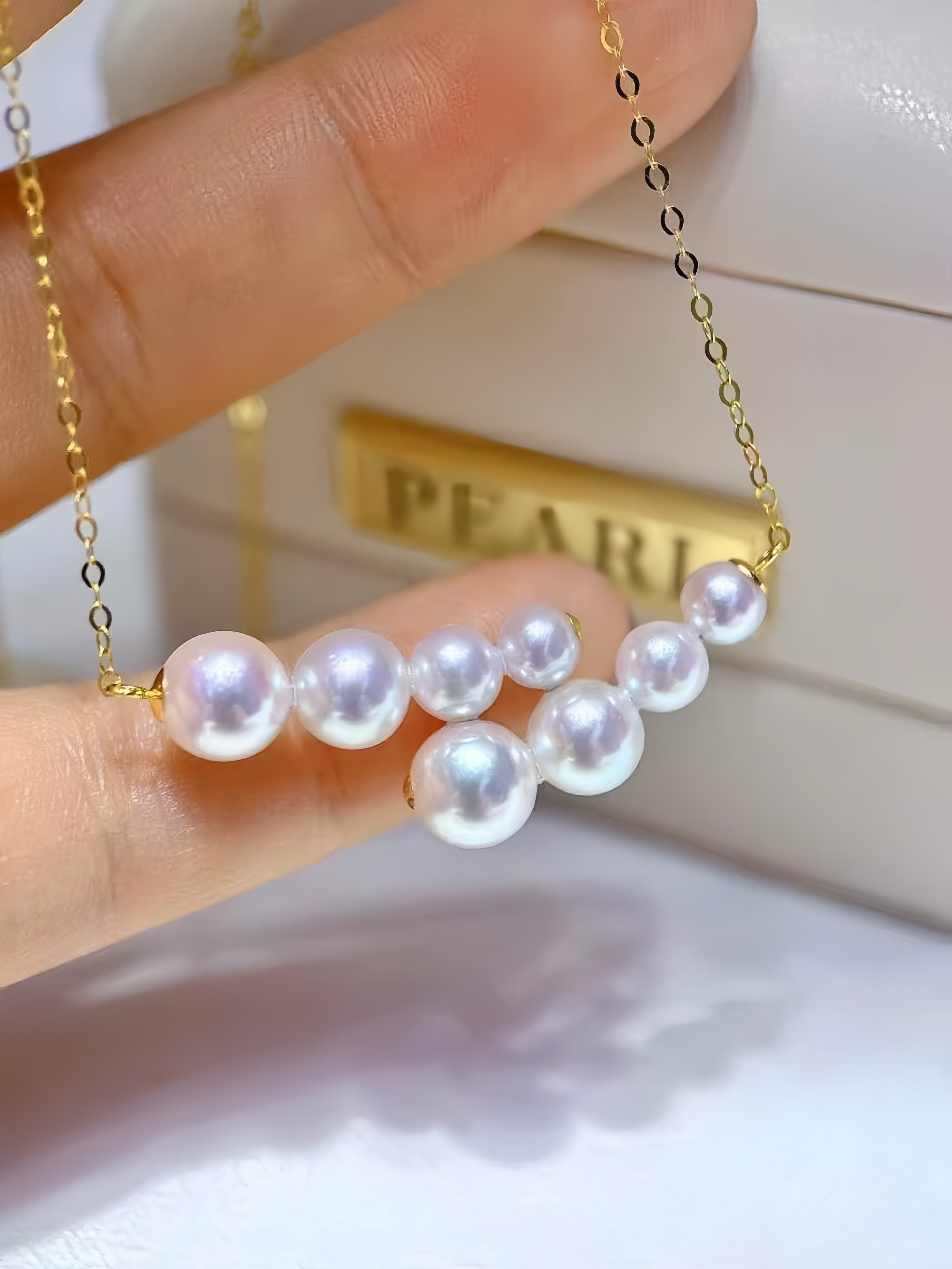 A close-up of a gold chain necklace with a row of white freshwater pearls draped across a person's fingers, with a white jewelry box in the background featuring the partial brand name 'PEARL'.