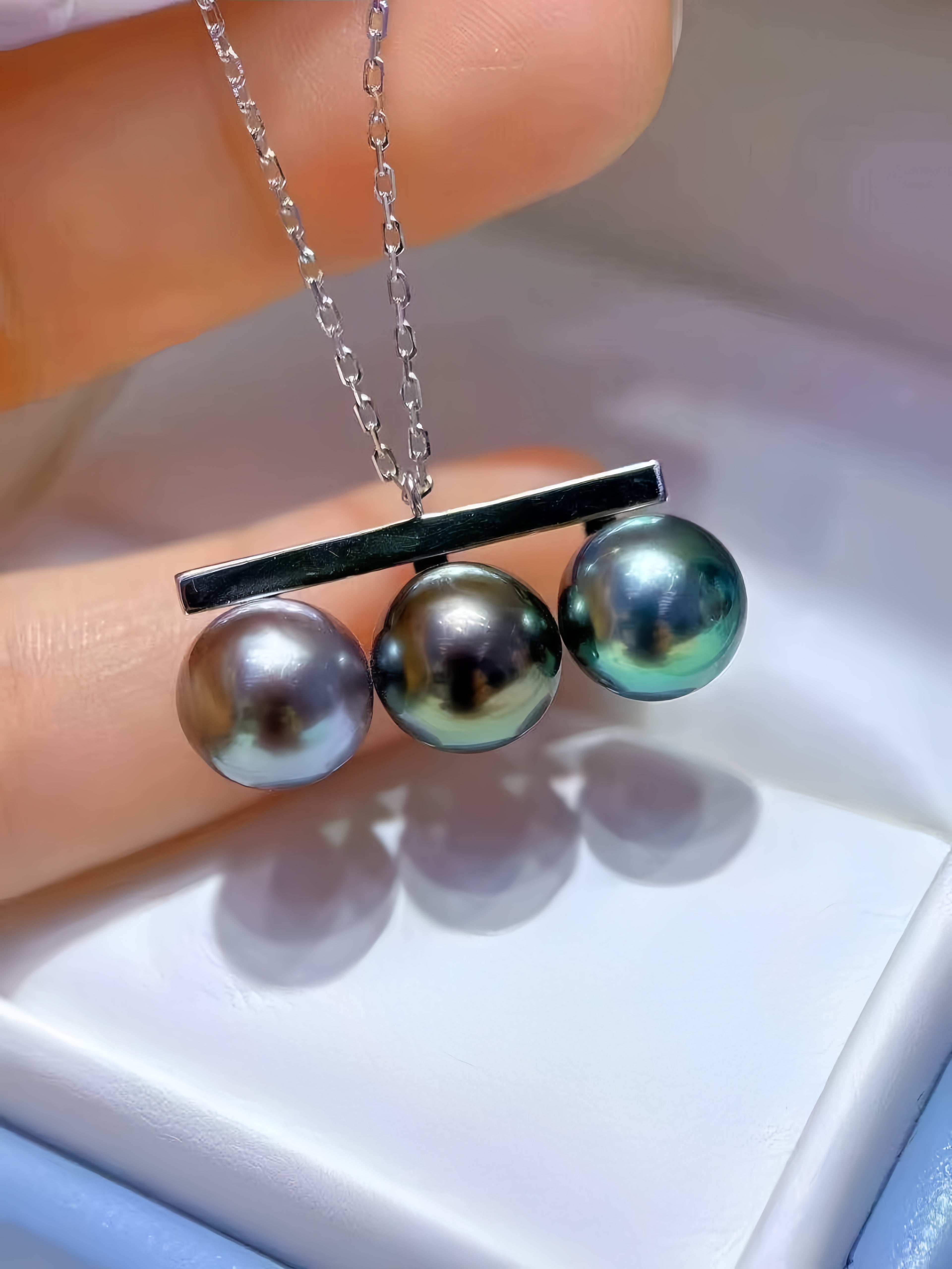 A close-up of a necklace with three round iridescent pearls suspended from a black bar on a silver chain, held between fingers over a white jewelry box.