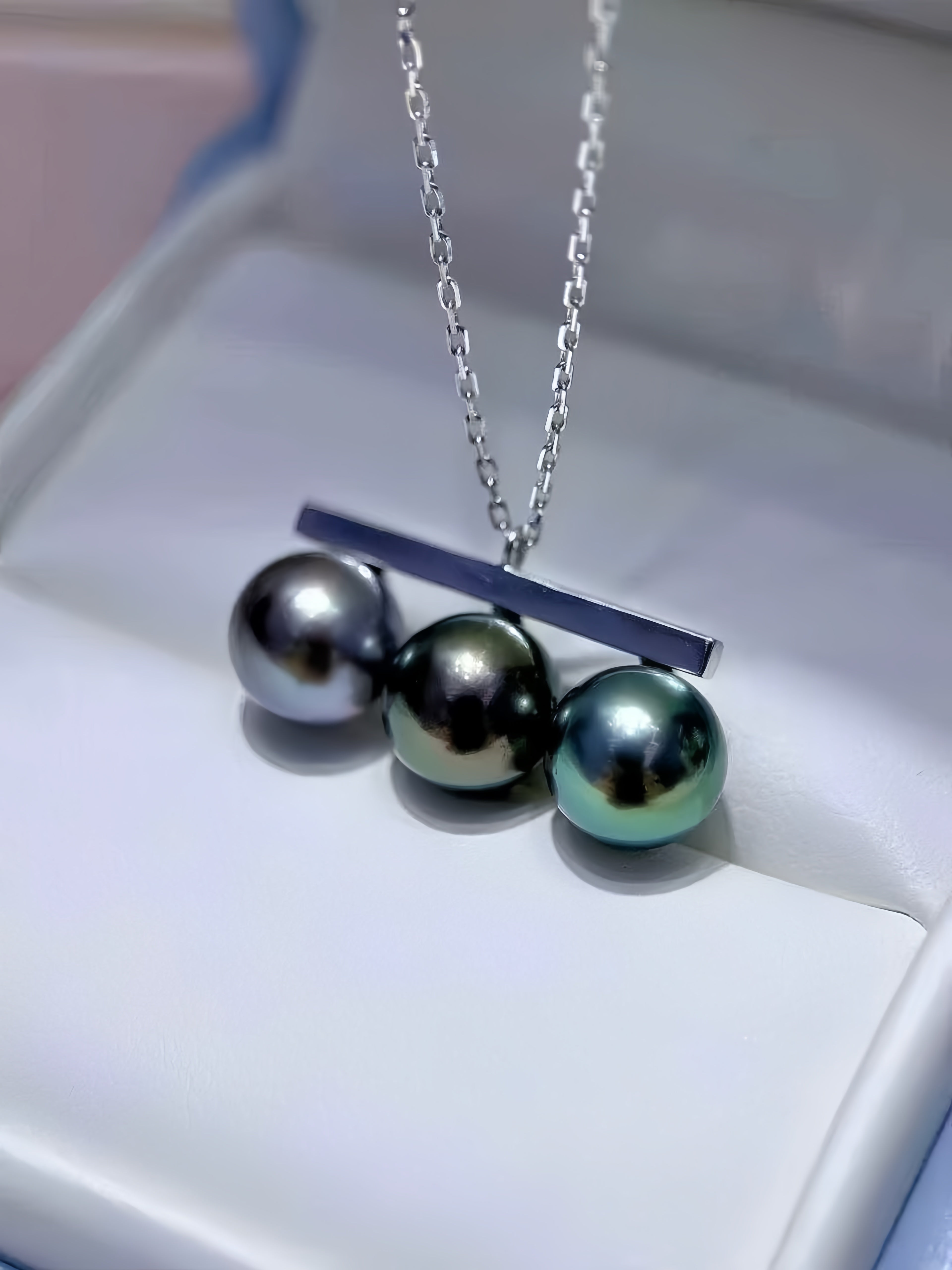 A silver pendant necklace featuring three large, iridescent freshwater pearls in gray, black, and green tones, suspended from a bar and displayed on a white jewelry box.