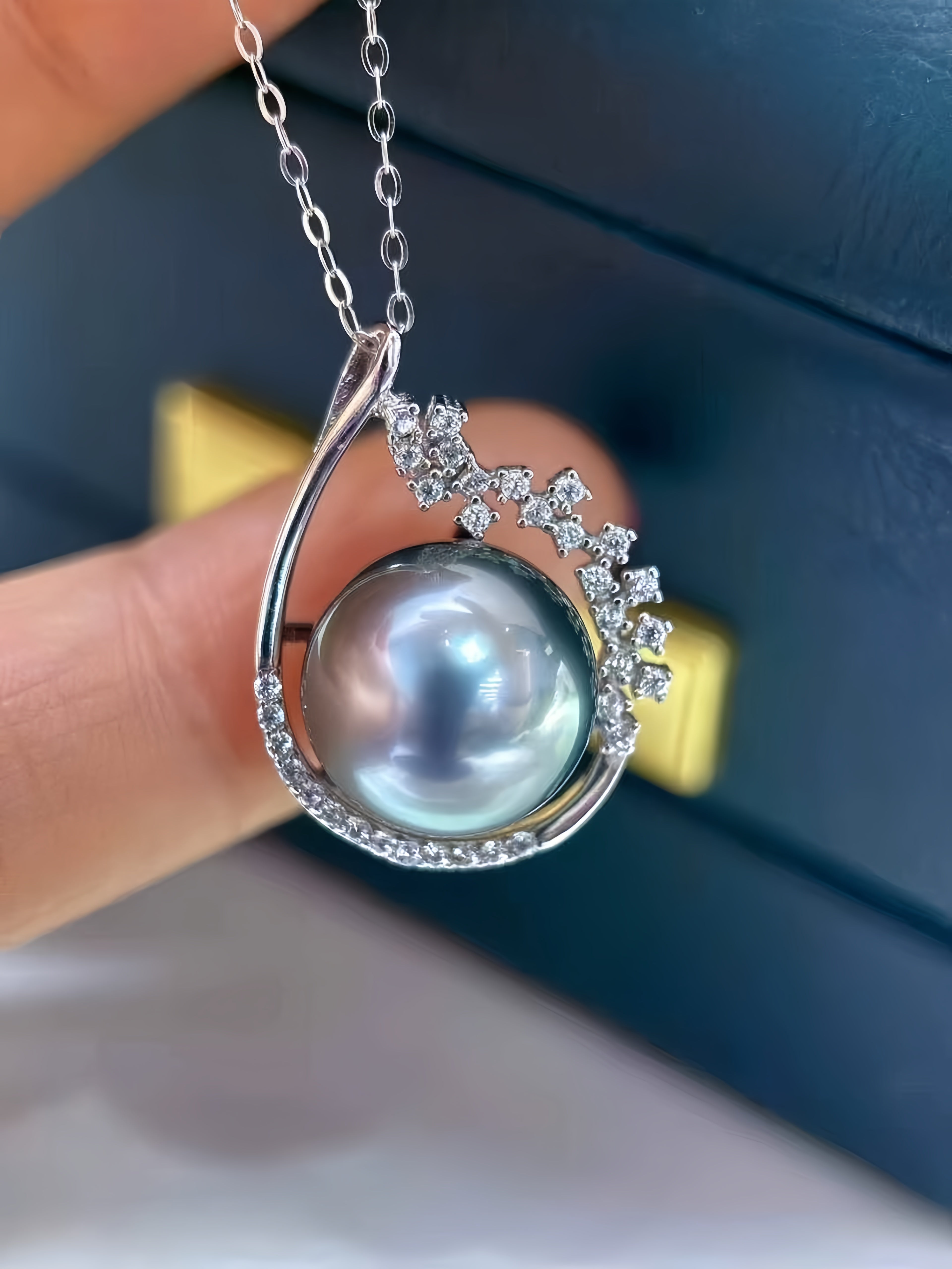 A close-up of a silver pendant necklace featuring a large round gray-blue pearl surrounded by small clear stones, held between fingers in front of a dark blue box.