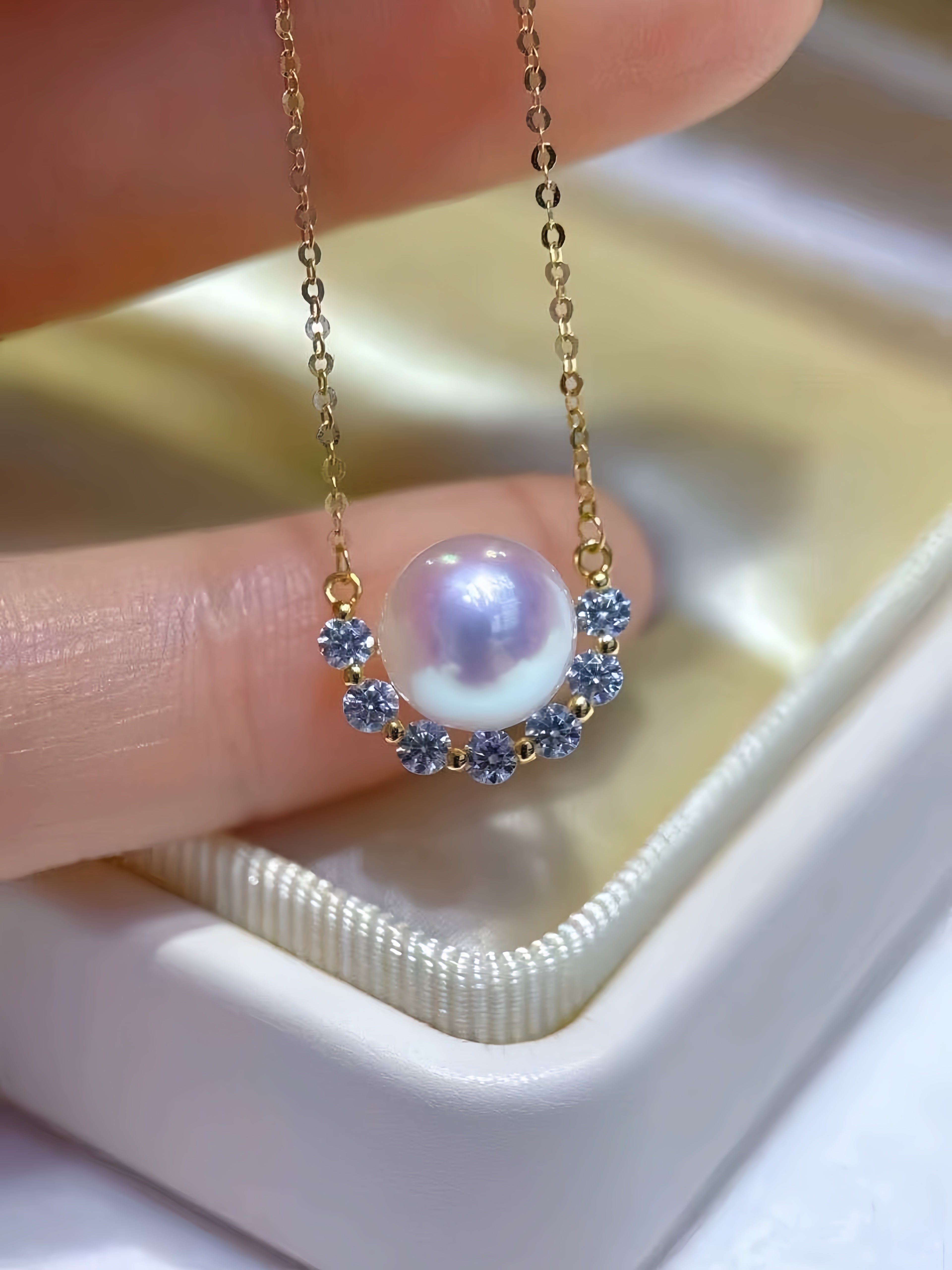 A close-up of a delicate gold necklace with a central lustrous pearl pendant, surrounded by small blue gemstones, held between fingers above a cream-colored jewelry box.