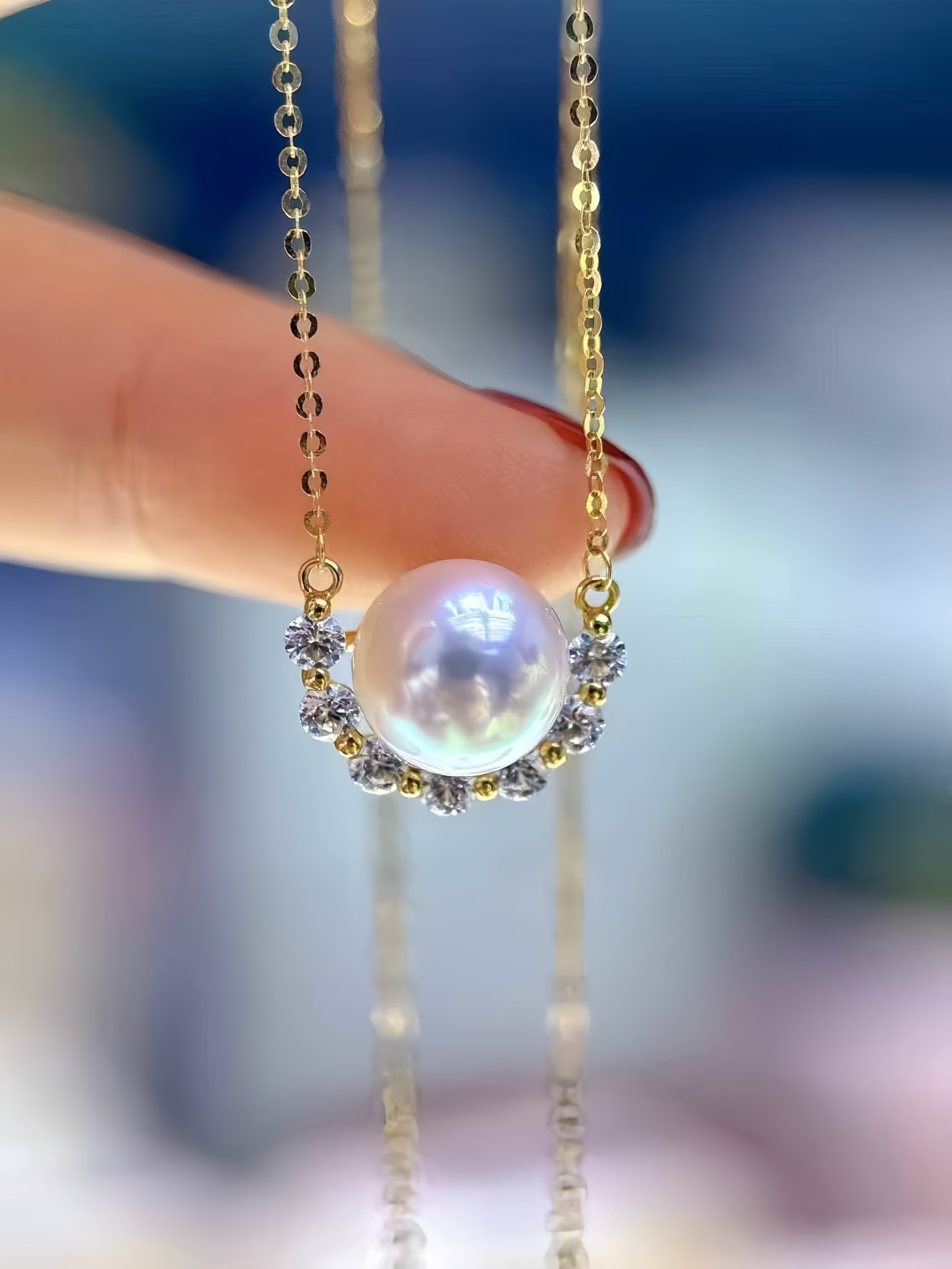 A close-up of a gold necklace with a large lustrous pearl pendant, held by a person's finger against a softly blurred blue background.