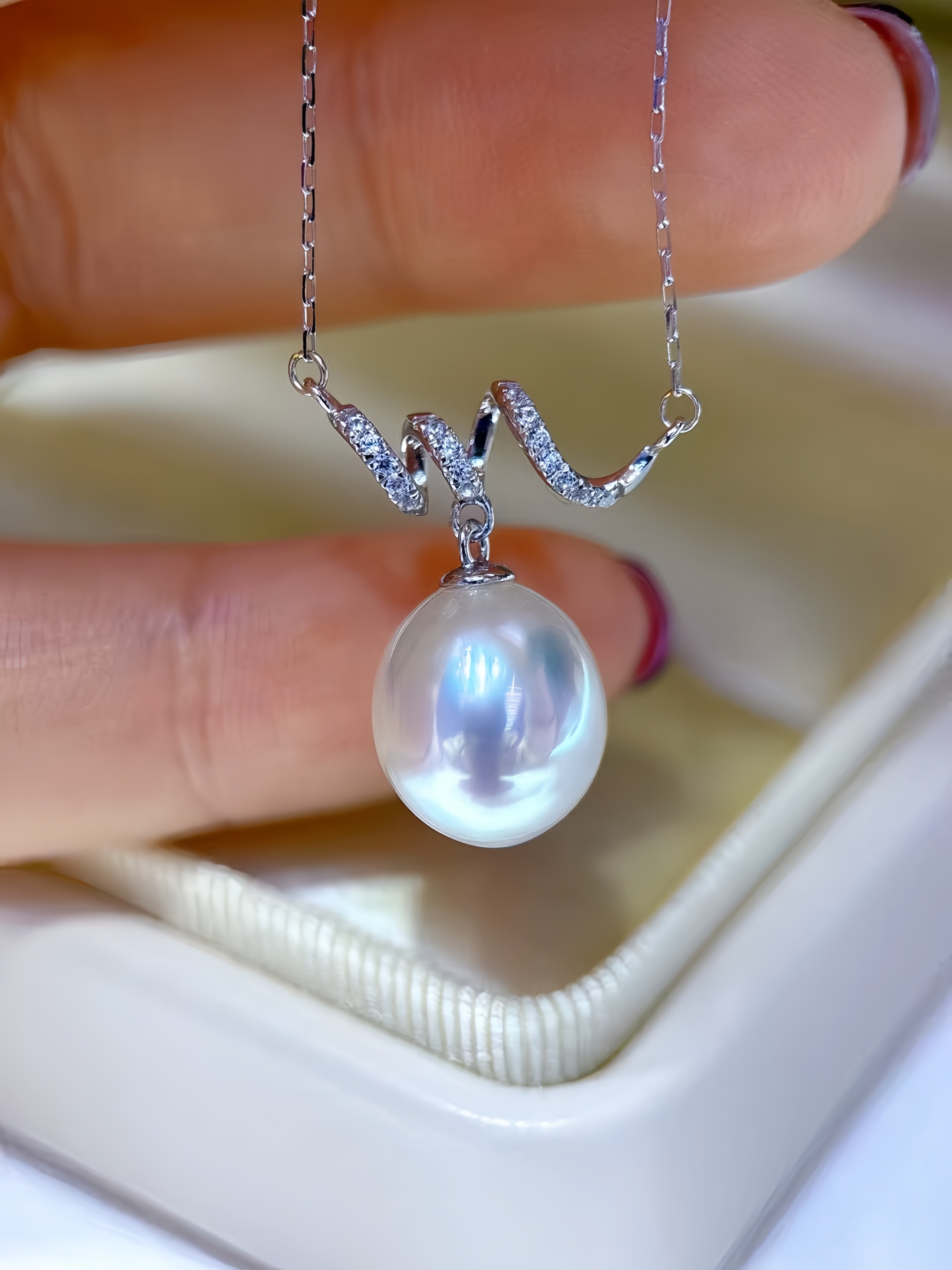 A close-up of a silver necklace with a large, lustrous white pearl pendant held by fingers above a cream-colored jewelry box.
