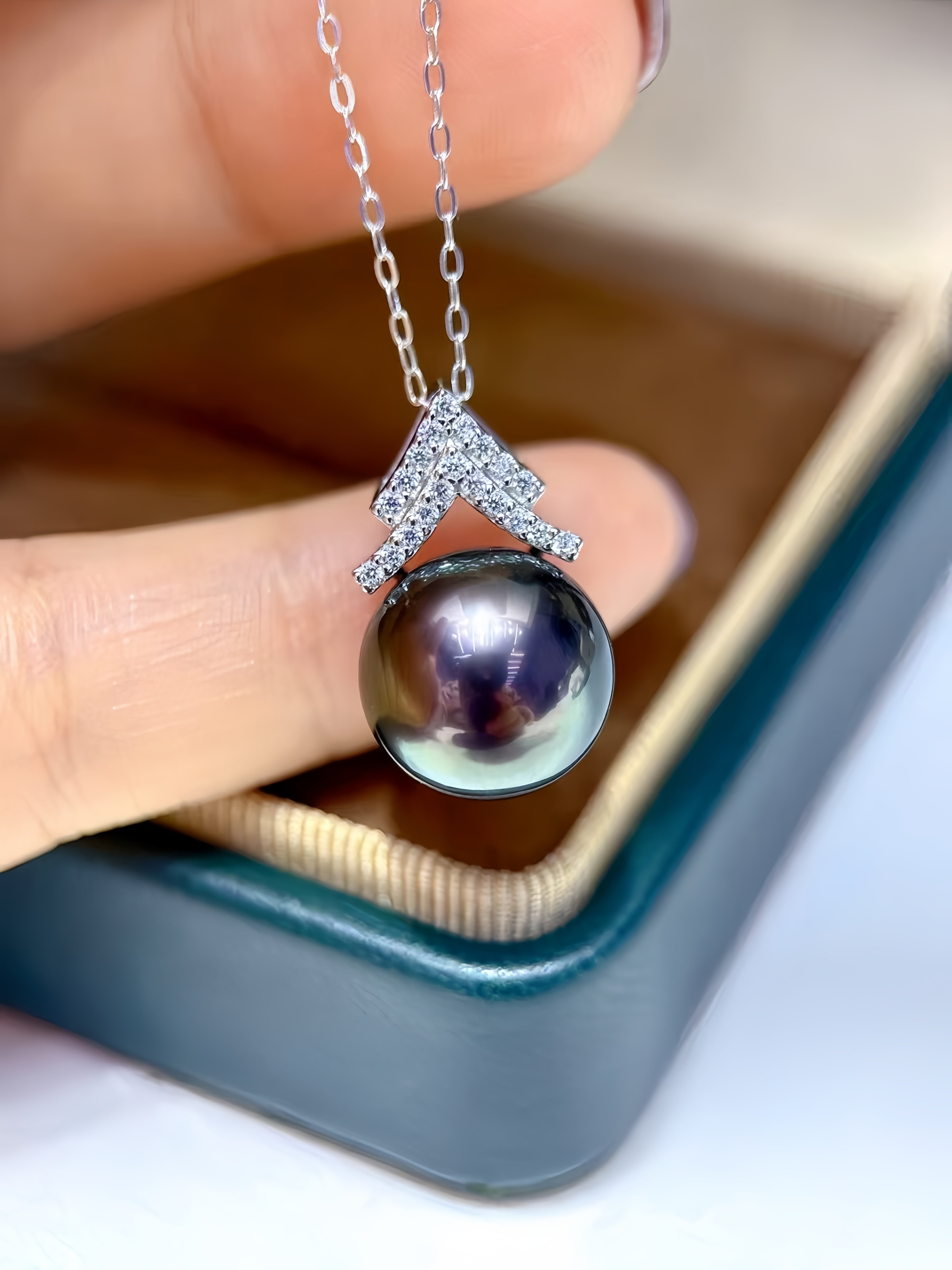 A close-up of a hand holding a silver pendant necklace with a large round dark pearl and a pavé-set chevron accent, positioned over an open jewelry box.