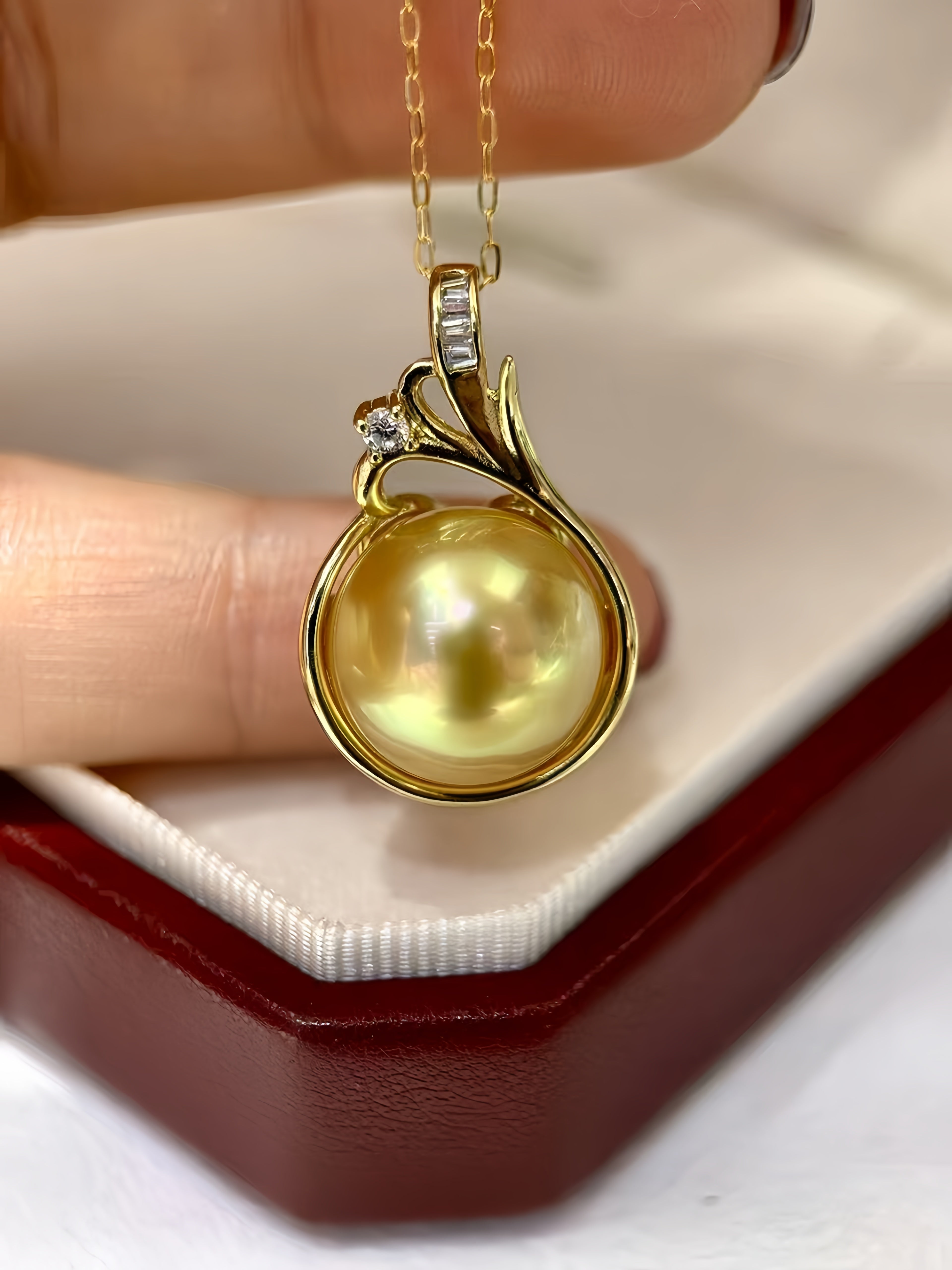 A gold pendant necklace with a large round yellow pearl and small clear stones, held between fingers above an open jewelry box.