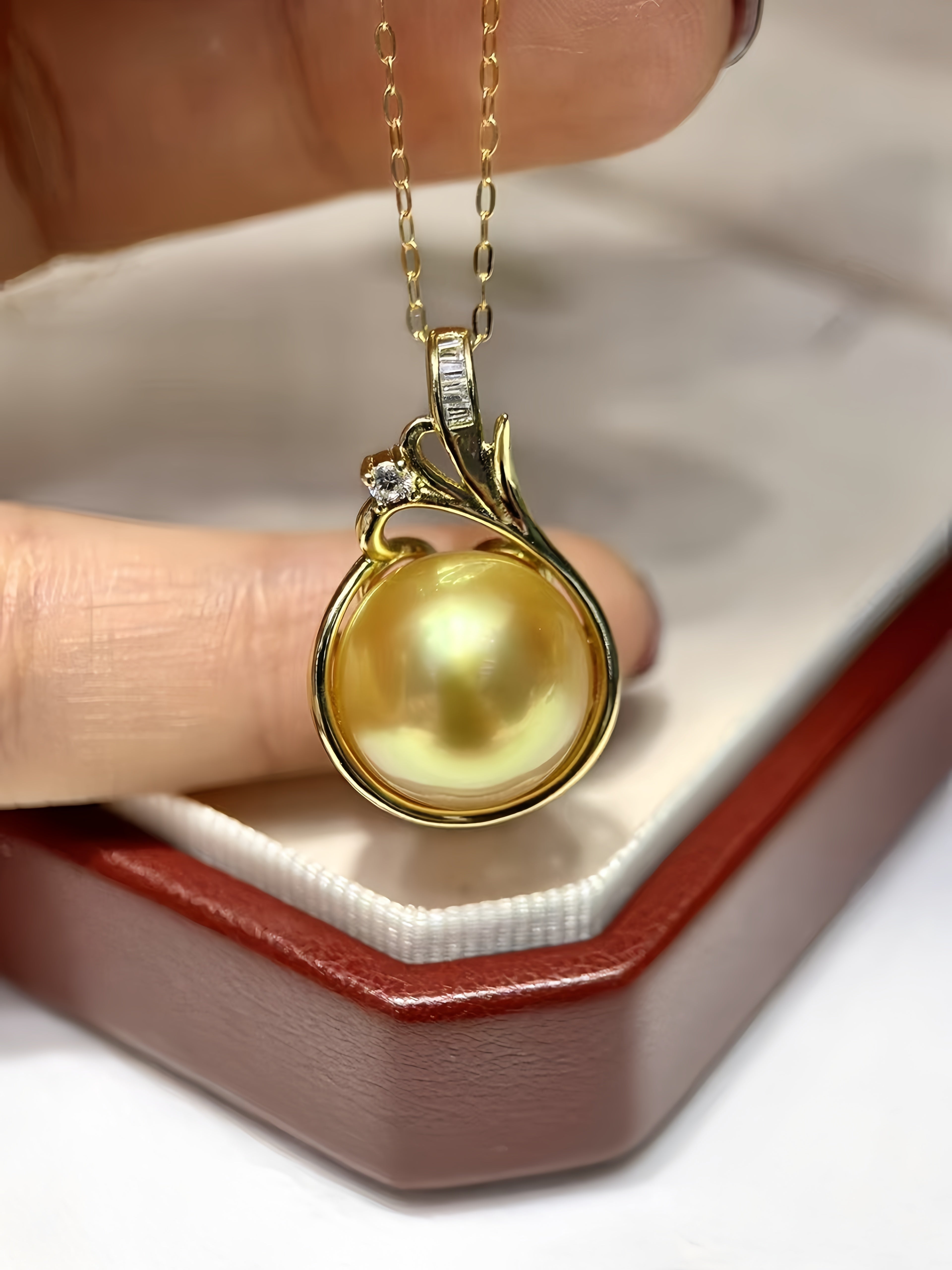 A gold pendant necklace with a large round yellow pearl and a small clear stone, held between fingers above an open jewelry box.