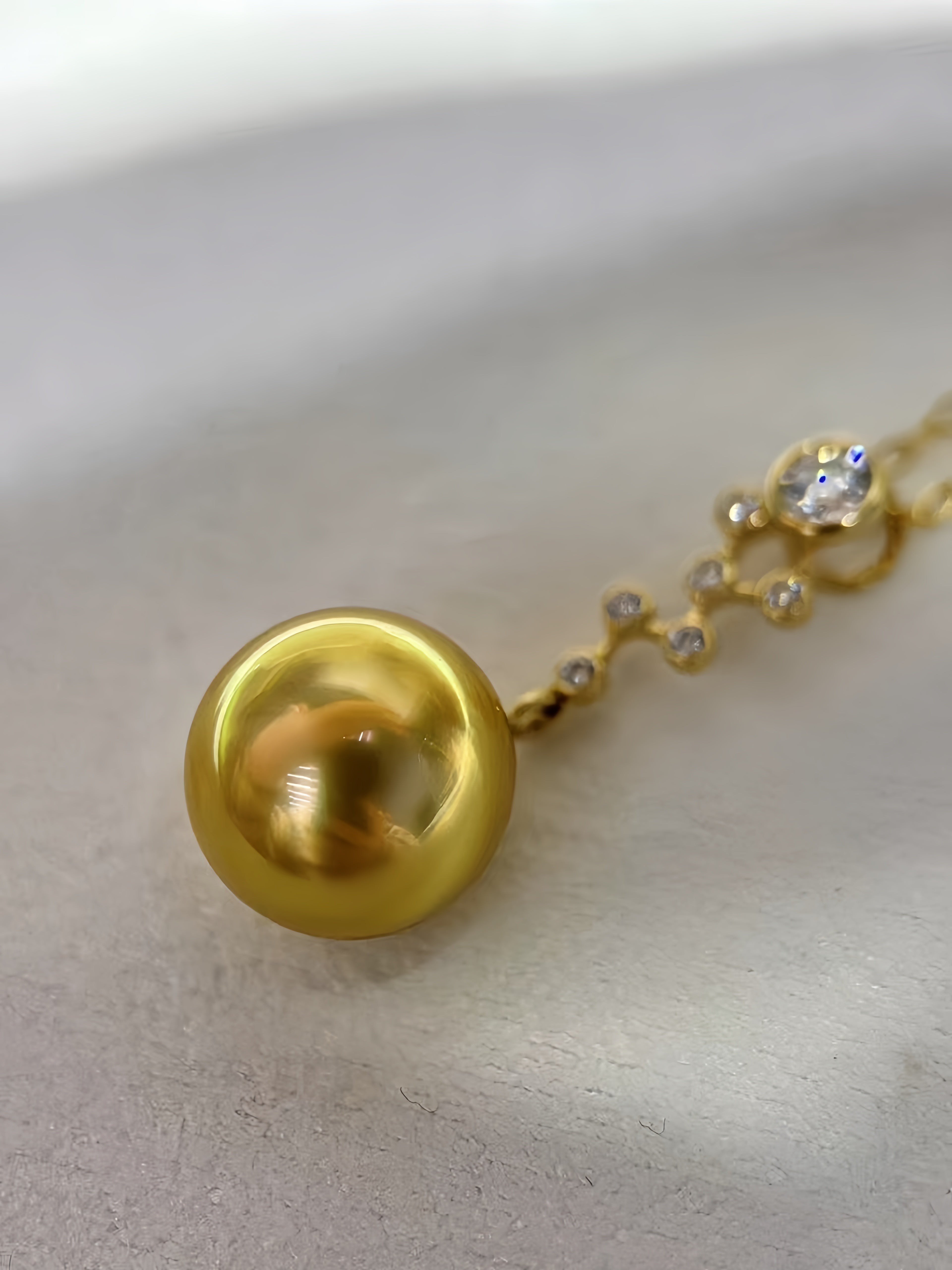 A close-up of a gold pendant necklace featuring a large, lustrous gold pearl pendant and a delicate gold chain with small clear stones, resting on a light beige textured surface.