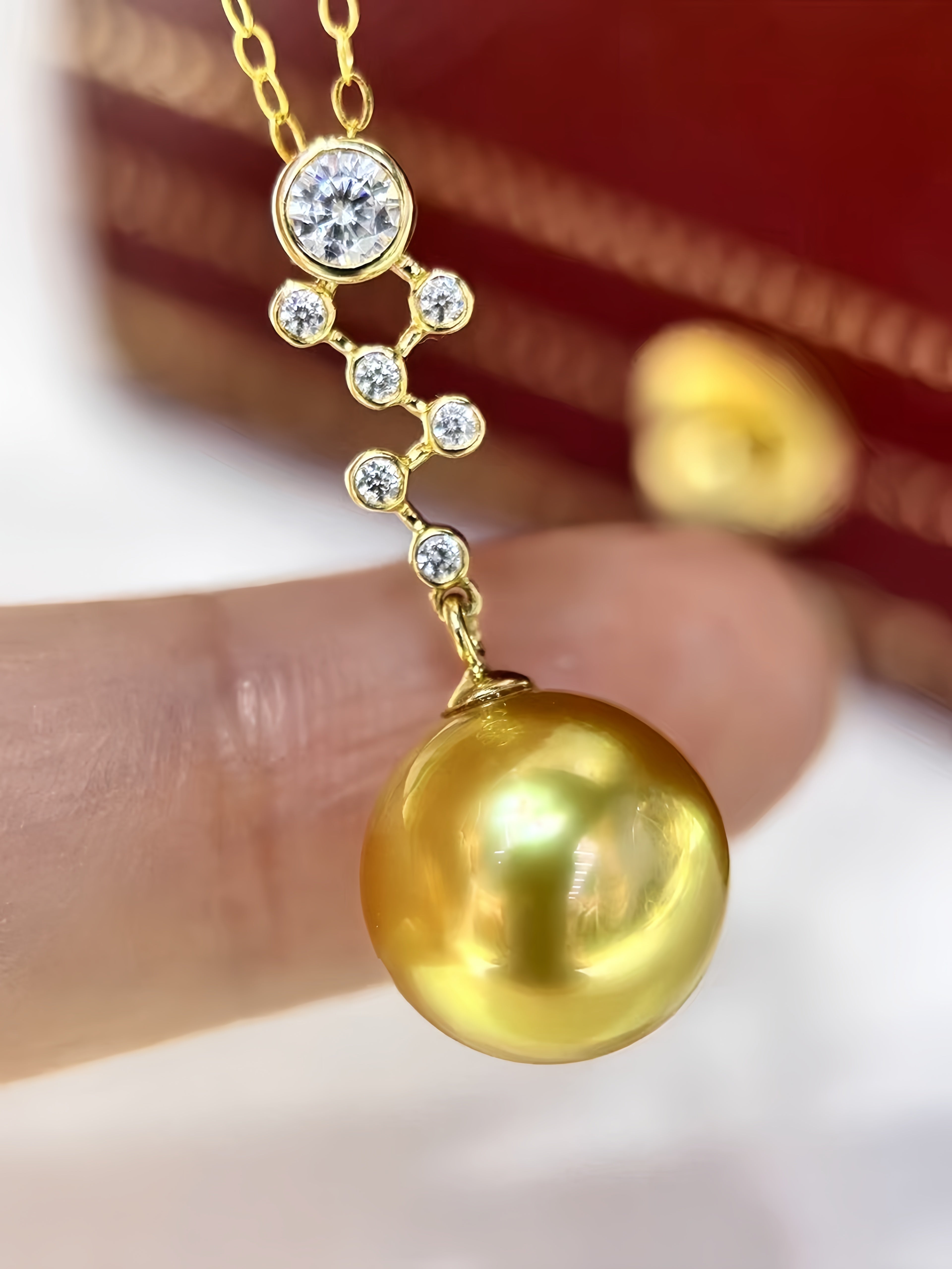 A gold pendant necklace with a large round yellow gold pearl and small clear stones, held between fingers against a blurred red and gold background.
