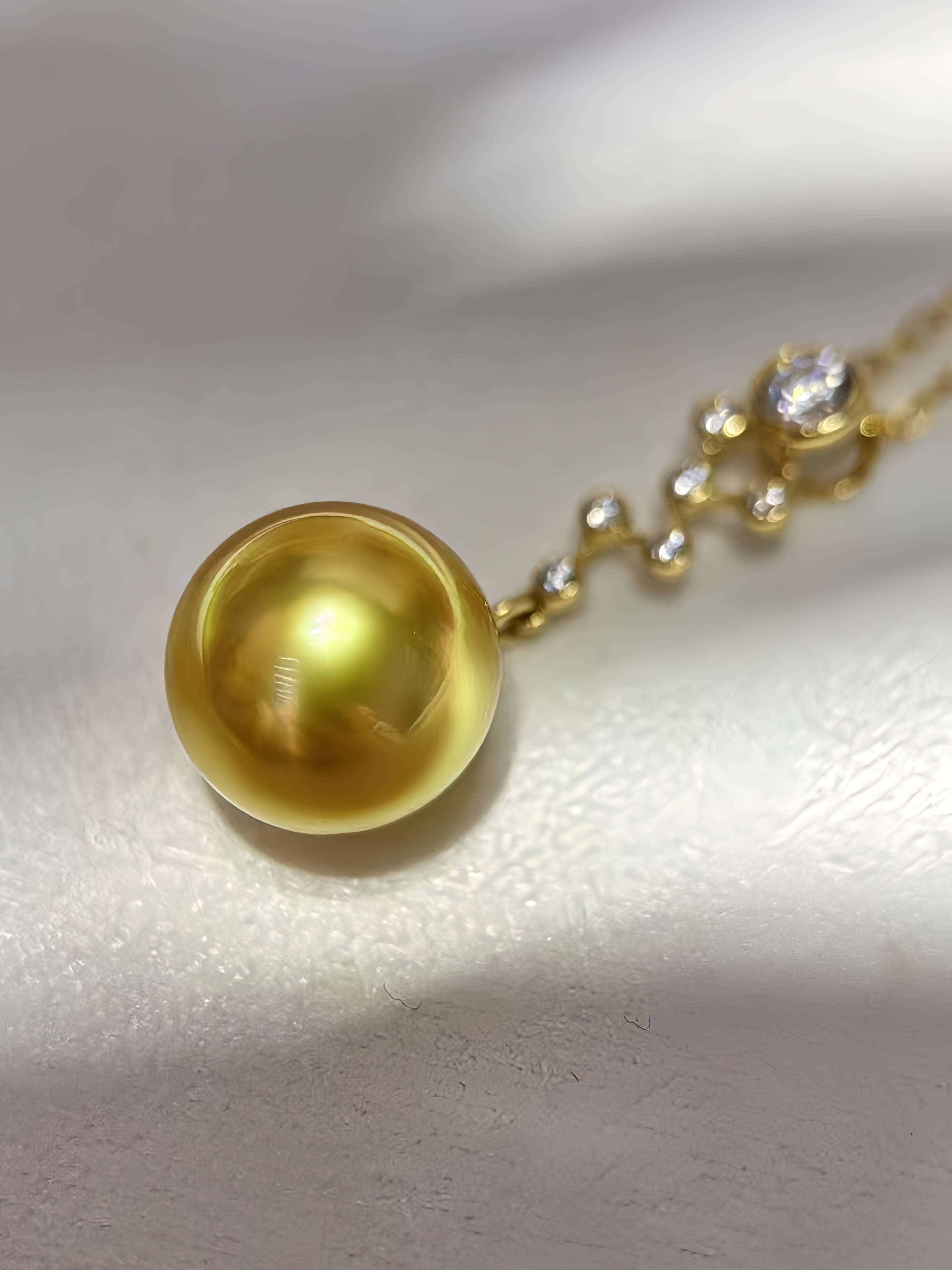 A close-up of a gold necklace featuring a large, round, lustrous yellow-gold pearl pendant, with a delicate gold chain and small clear stones on a textured white surface.