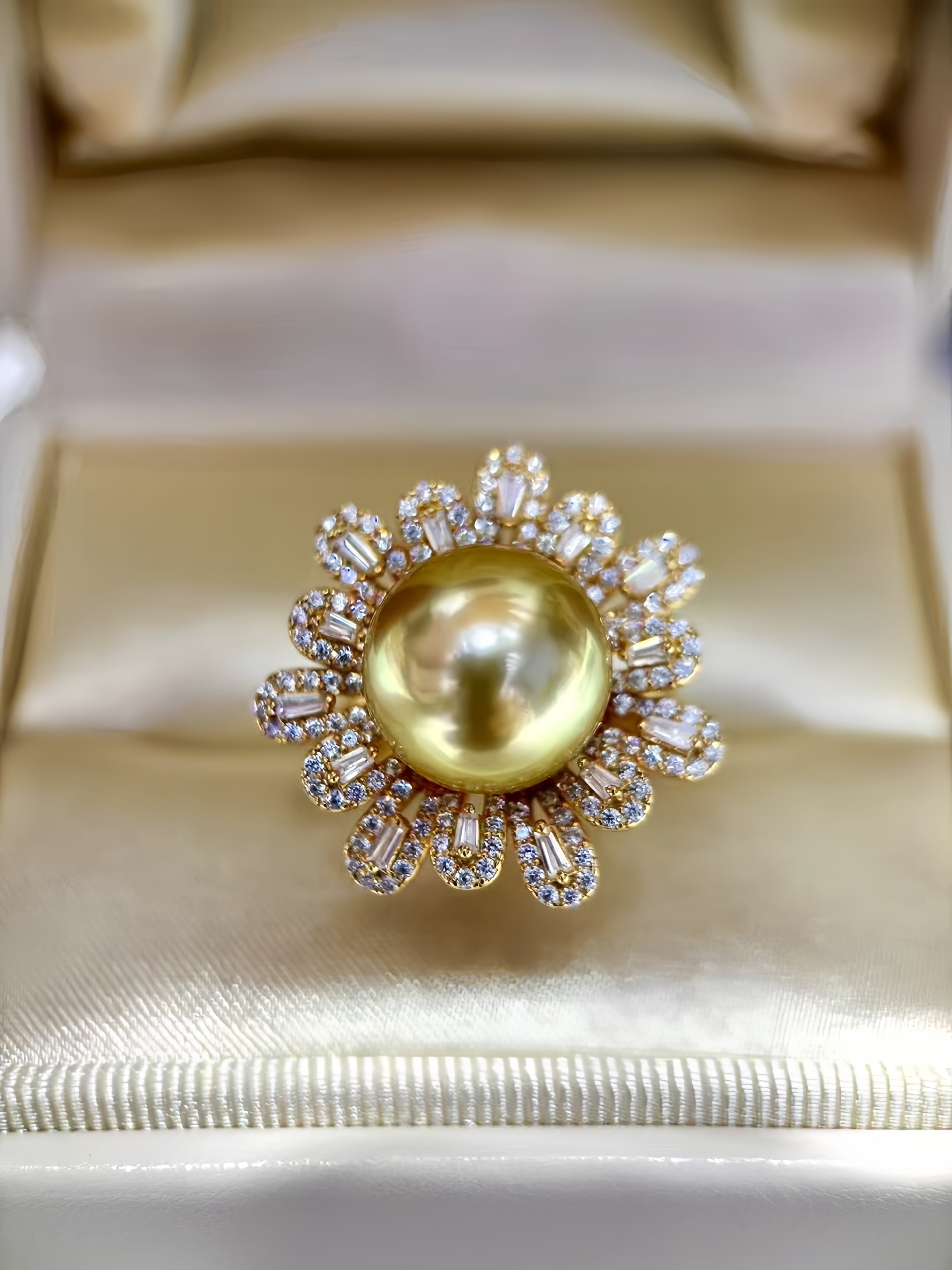 A gold ring featuring a large, lustrous yellow pearl surrounded by a halo of small clear stones, displayed on a cream satin ring box.
