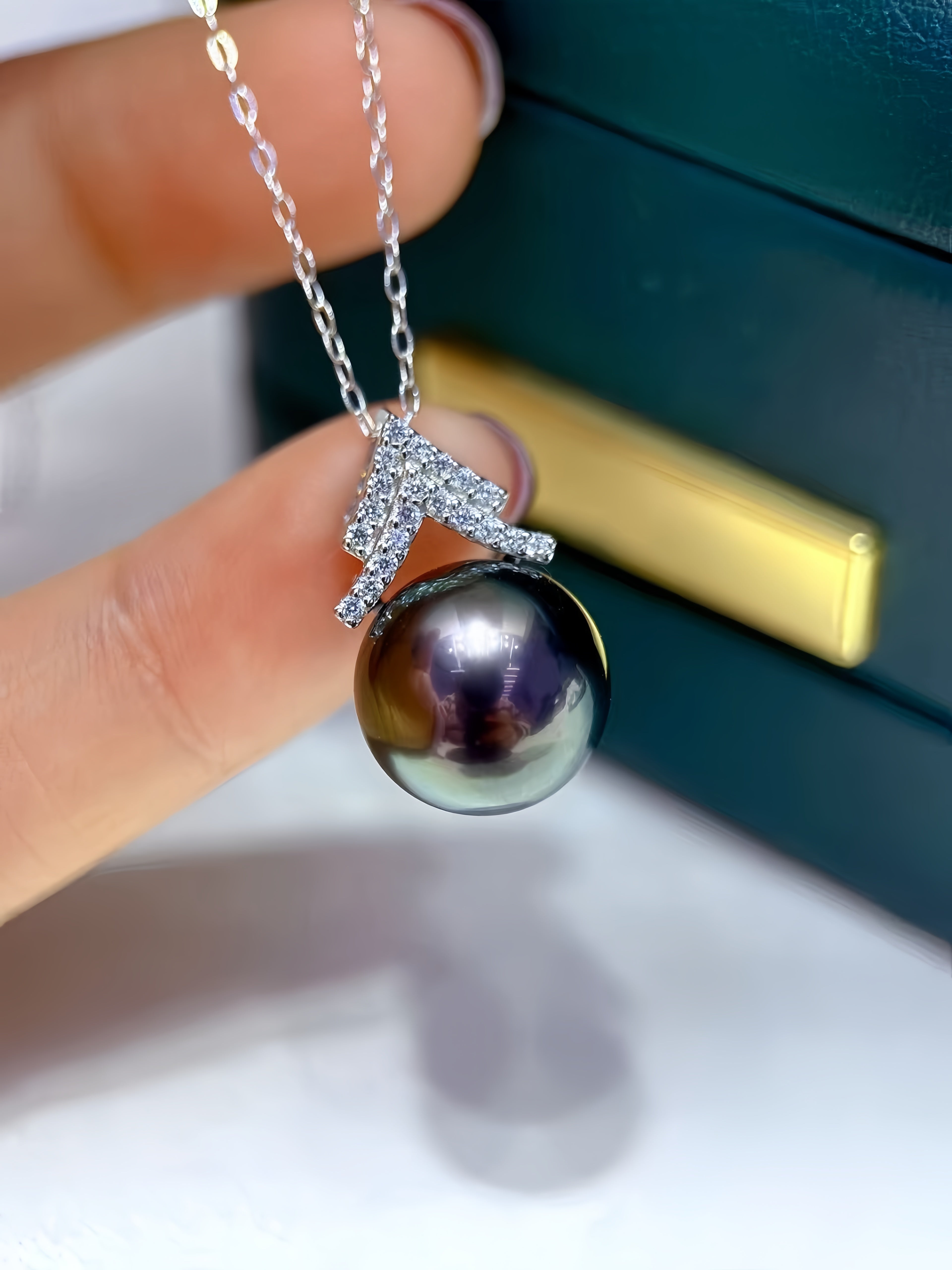 A close-up of a hand holding a silver pendant necklace with a large, dark, iridescent pearl and a silver mountain-shaped setting adorned with small clear stones, with a dark green jewelry box in the background.