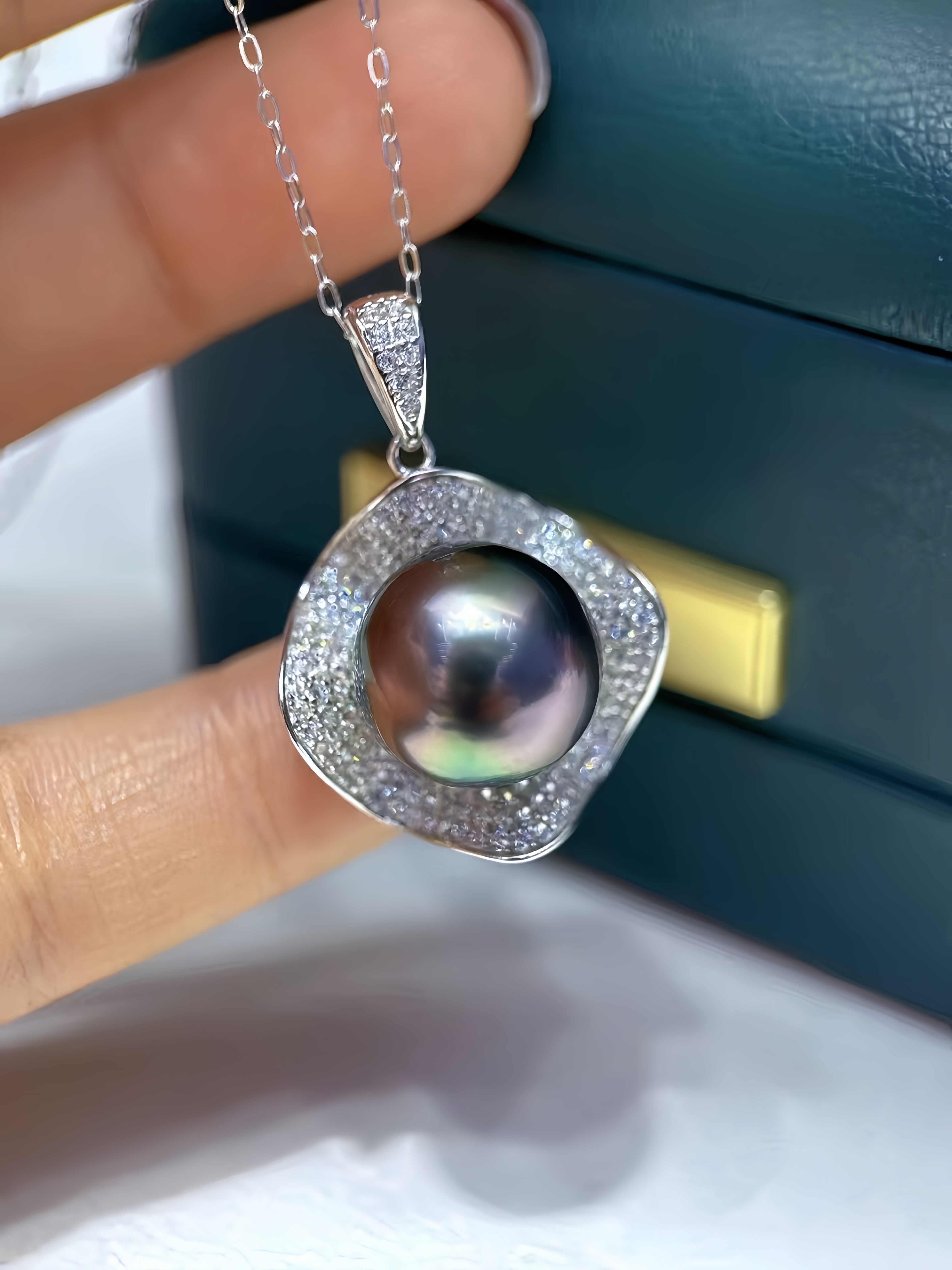 A close-up of a hand holding a silver pendant necklace with a large iridescent pearl surrounded by a halo of small clear stones, with a dark teal jewelry box in the background.