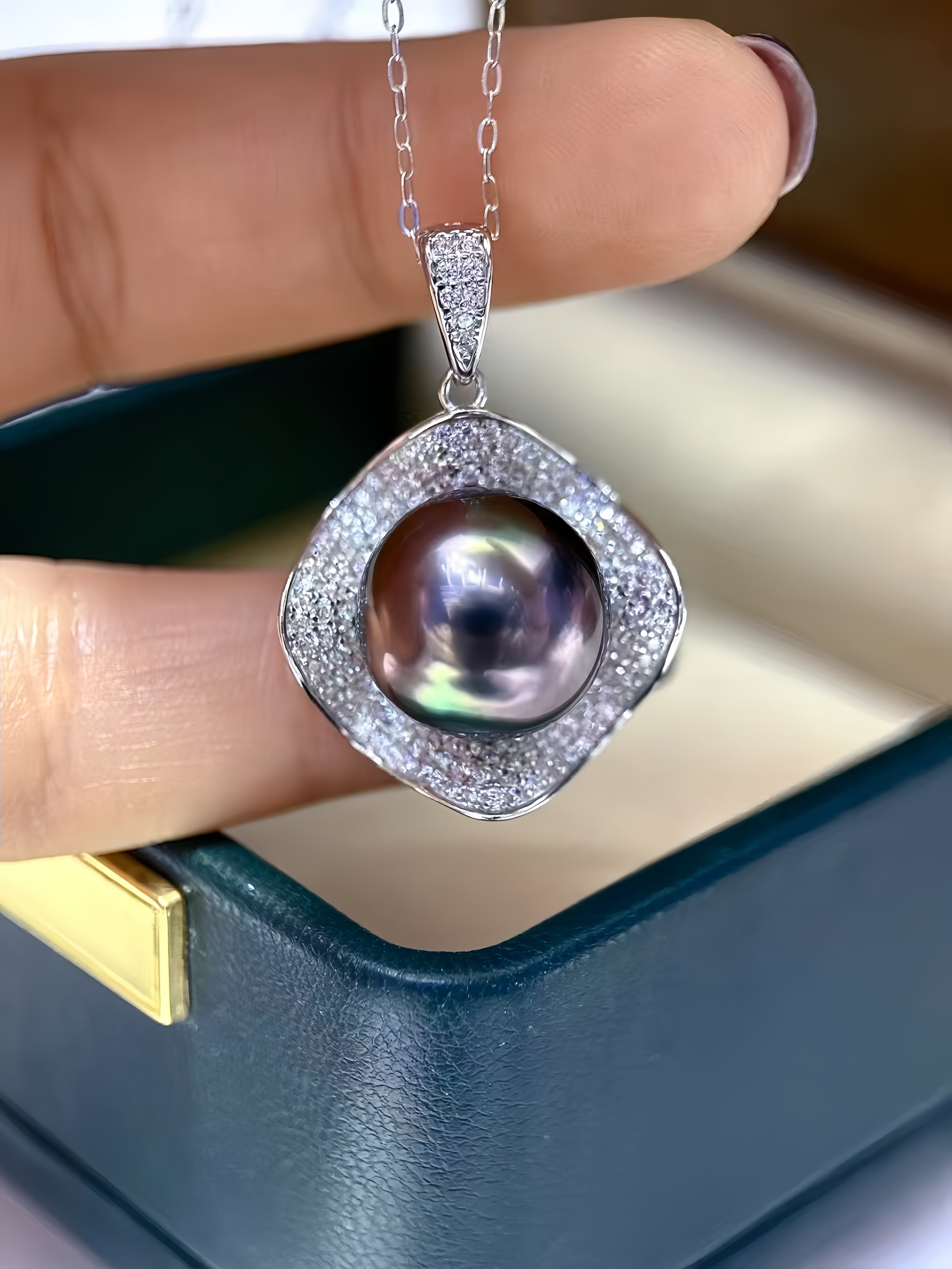 A close-up of a hand holding a silver pendant necklace with a large iridescent pearl surrounded by a halo of small clear stones, positioned above an open dark teal jewelry box.