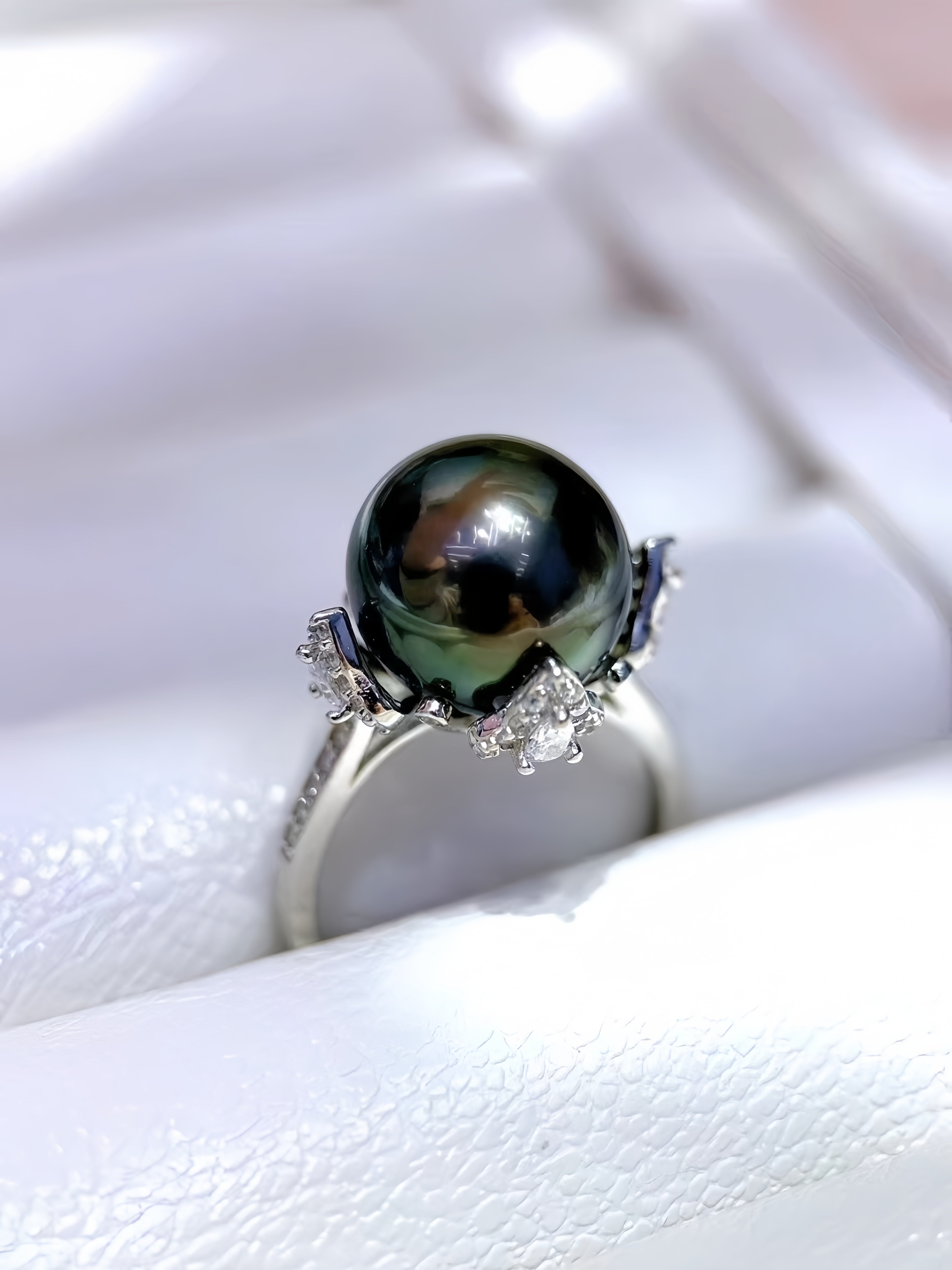 A close-up shot of a ring featuring a large, round, dark green-black Tahitian pearl set in a silver-colored metal with small clear accent stones, resting on a white textured ring box.