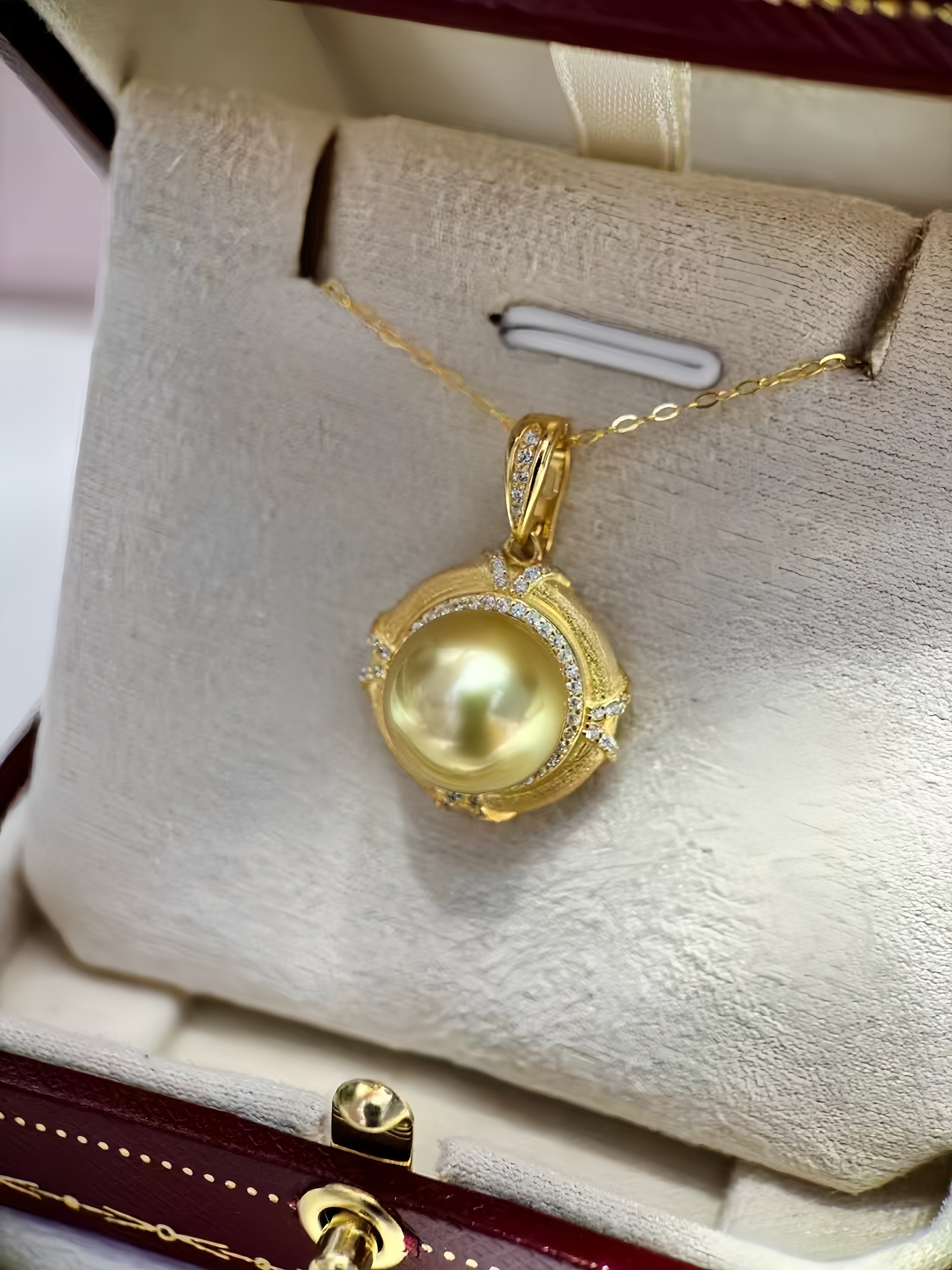 A gold pendant necklace with a large round yellow pearl and small clear stones, displayed inside a cream-colored jewelry box.
