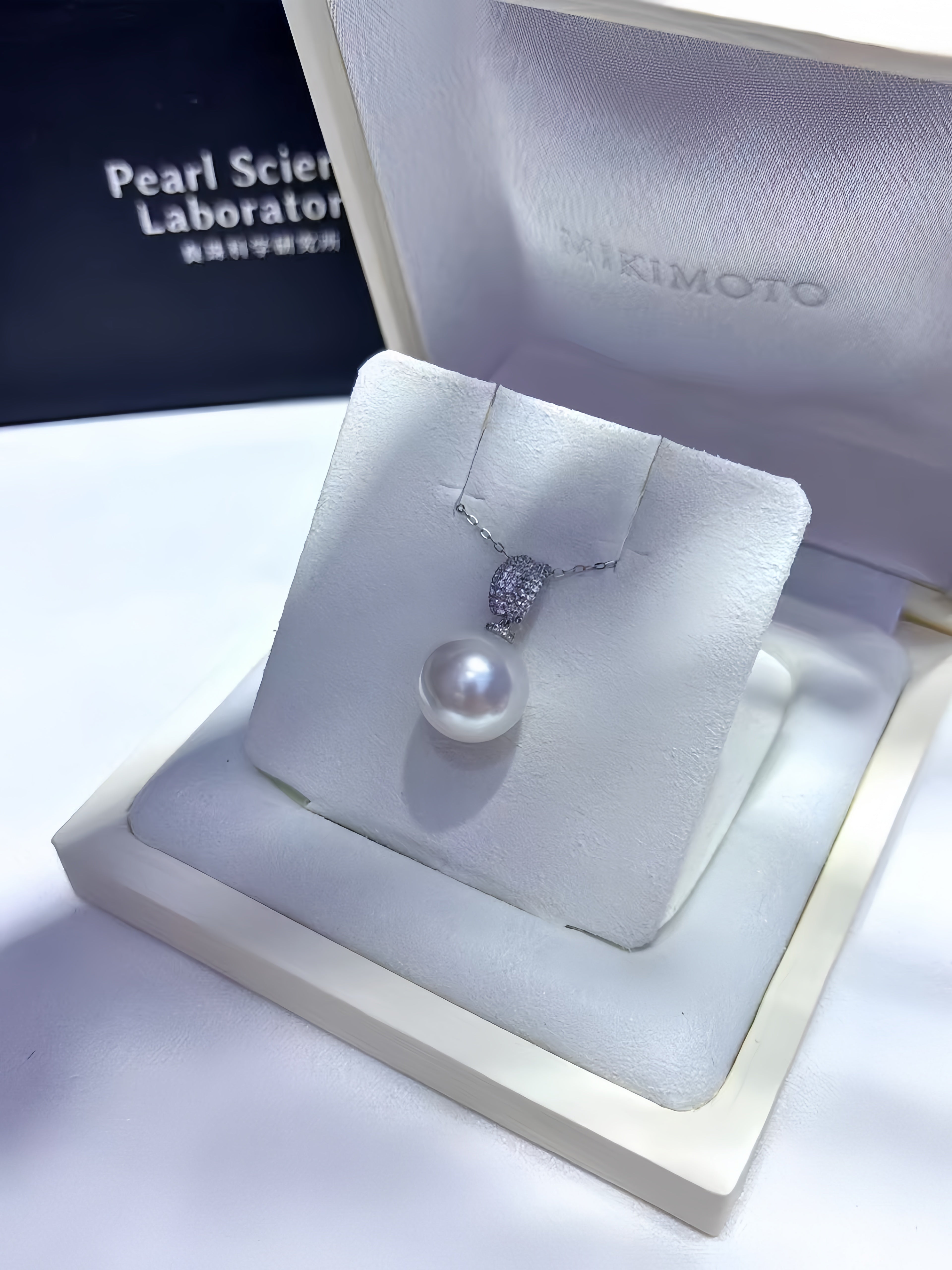 A pearl pendant necklace displayed on a white jewelry stand inside an open presentation box. The pendant has a white pearl with a silver-toned setting, and the box interior shows the brand name Takimoto.