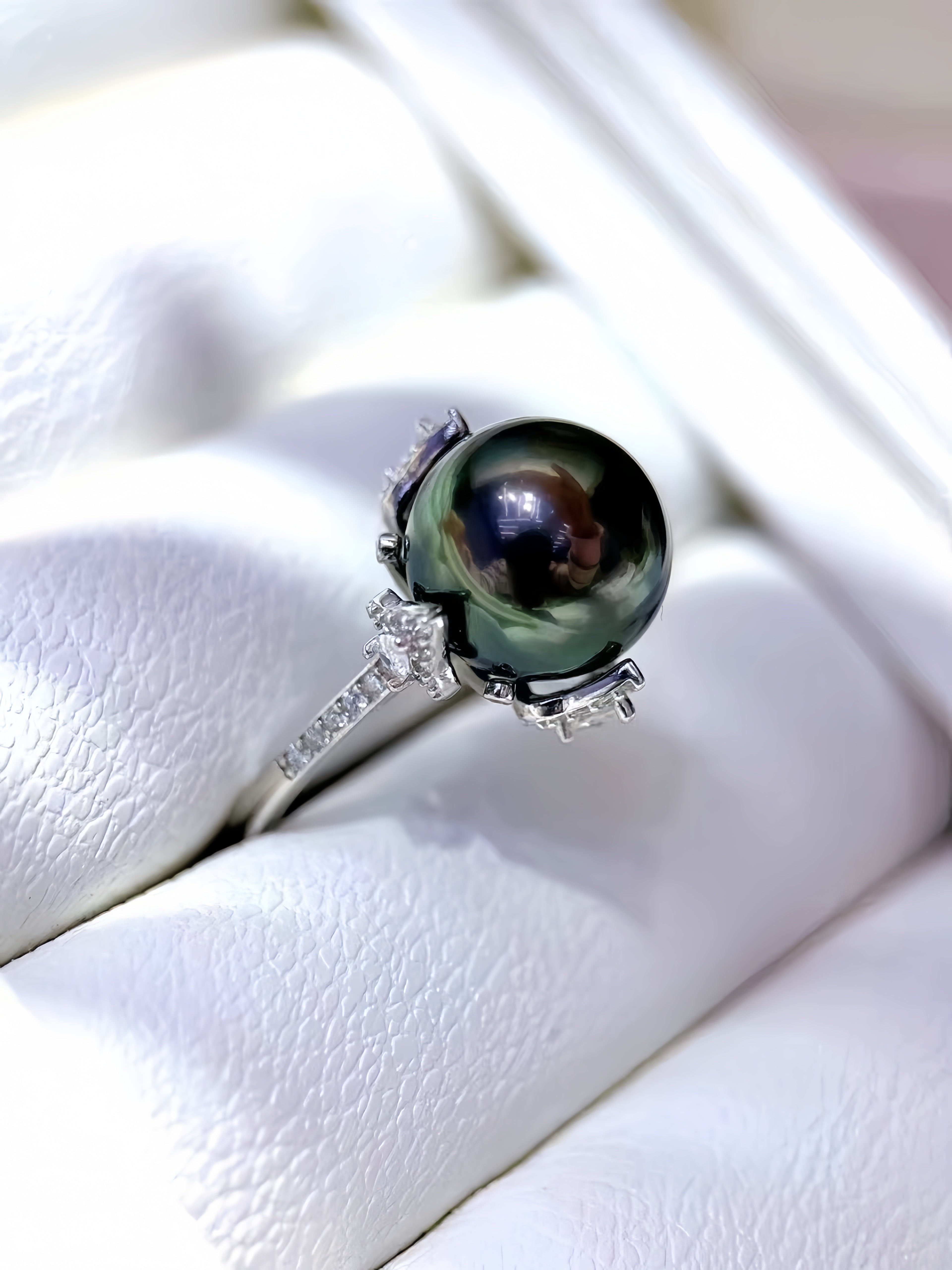 A close-up of a ring featuring a large round dark pearl in a silver-toned setting with small clear stones on the band, displayed on a white textured cushion.