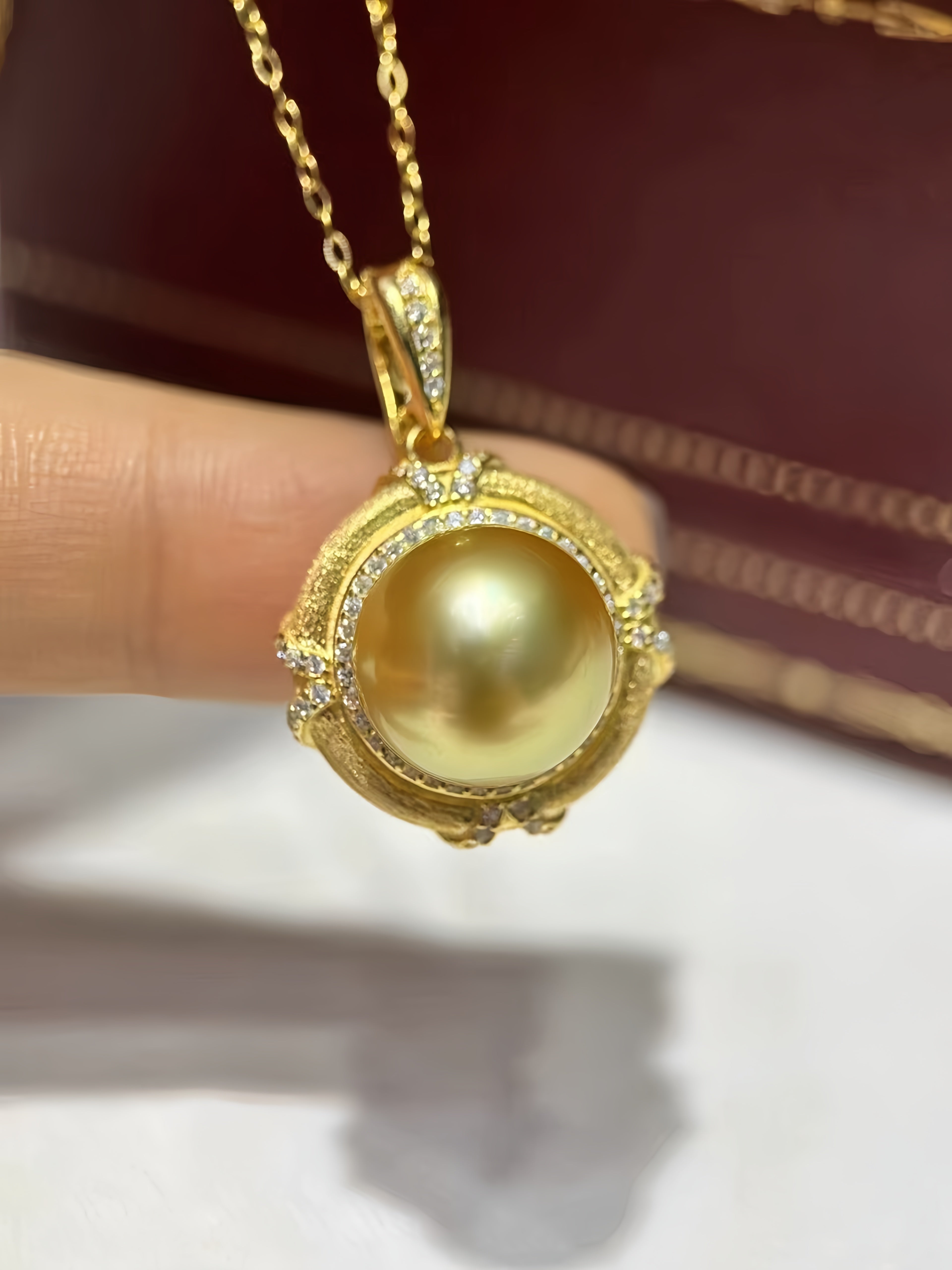 A gold pendant necklace featuring a large round yellow pearl surrounded by small clear stones, held by a person's fingers against a dark red box and white surface background.