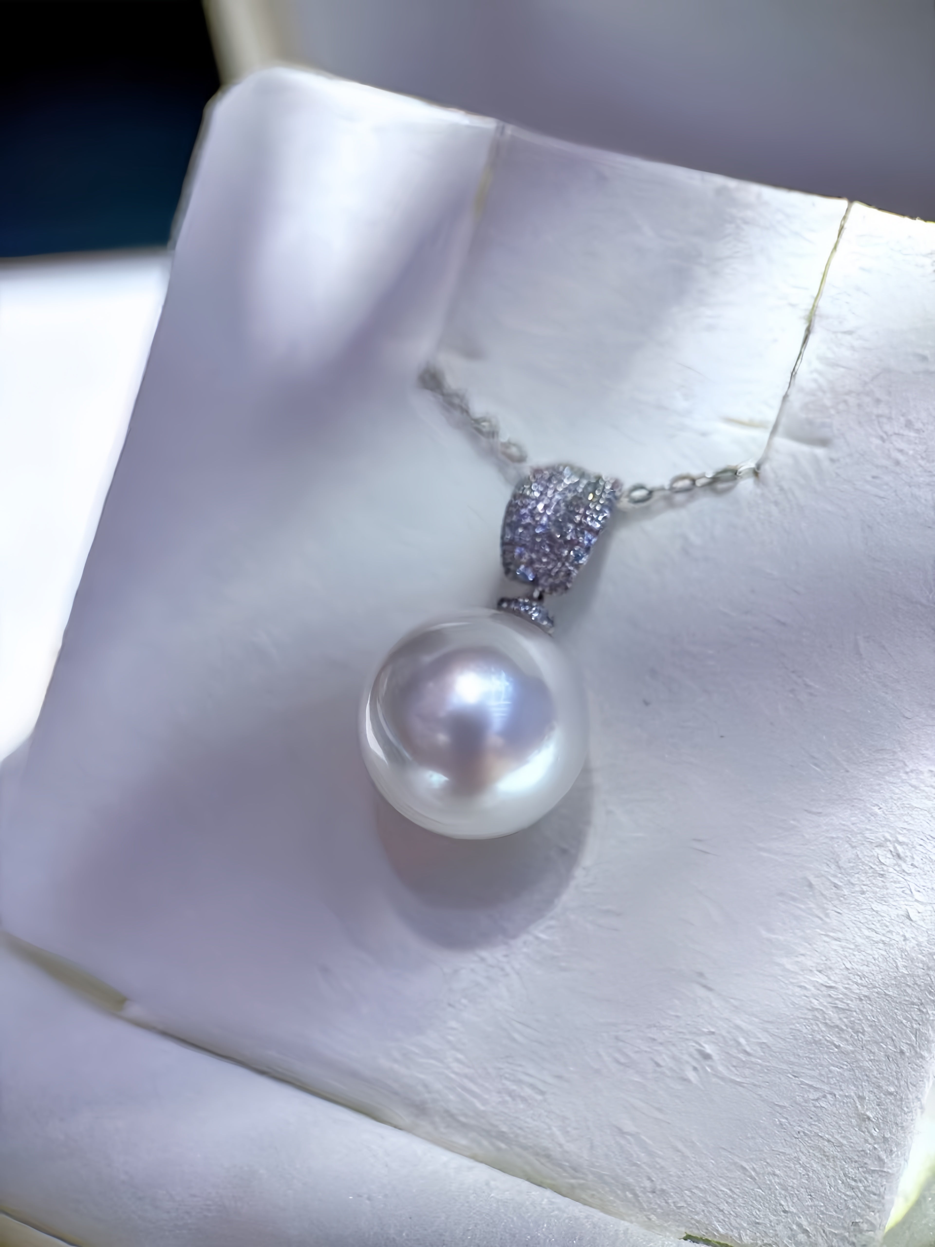 A silver pendant necklace featuring a single lustrous white pearl and a pavé-set silver bail, displayed on a white jewelry box cushion.