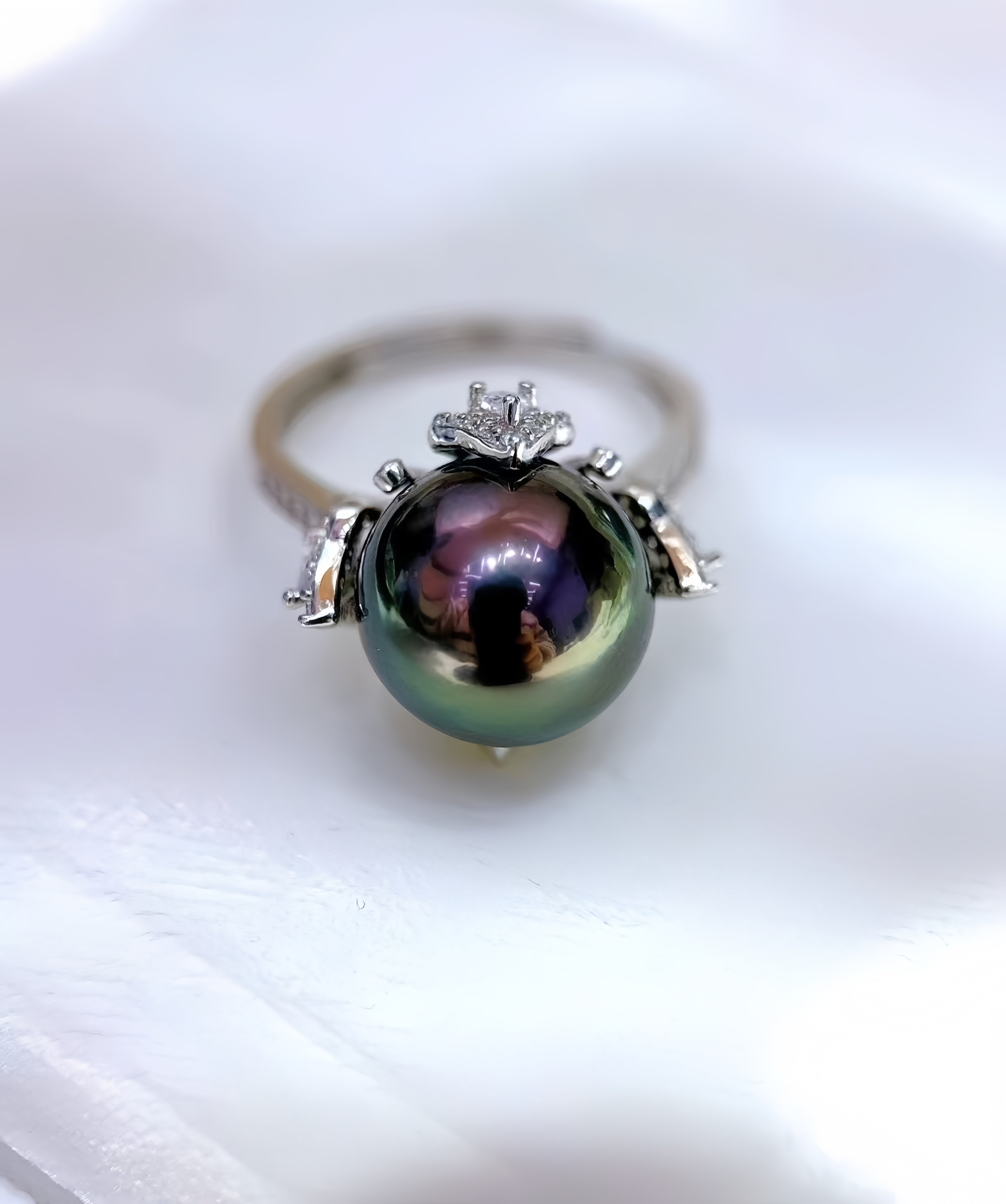 A silver ring featuring a large, round, iridescent green-black pearl with small clear accent stones, resting on a soft white fabric background.