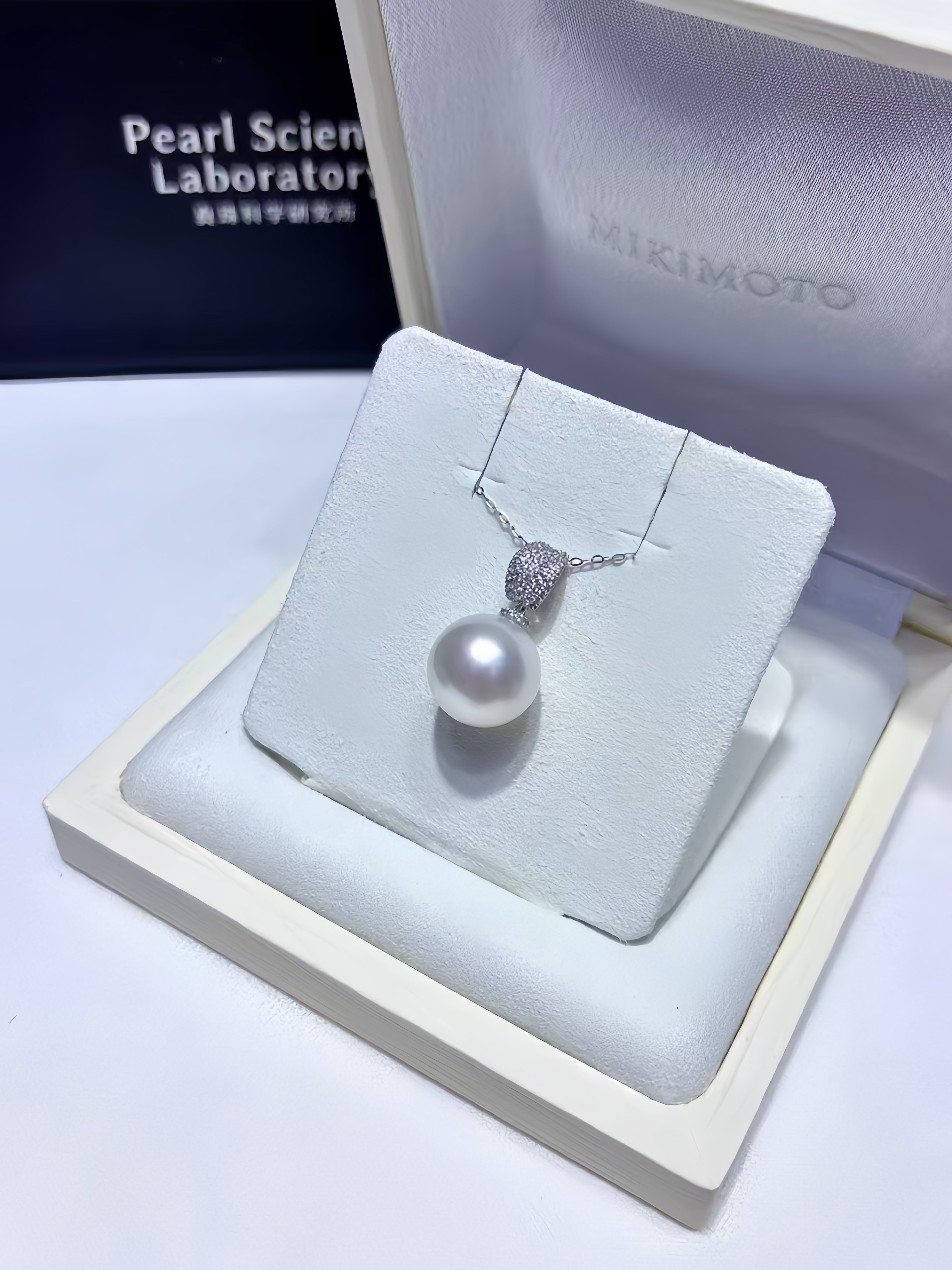 A pearl pendant necklace displayed on a white jewelry stand inside an open Mikimoto box. The necklace features a large white pearl hanging from a silver-toned chain with a textured silver clasp.