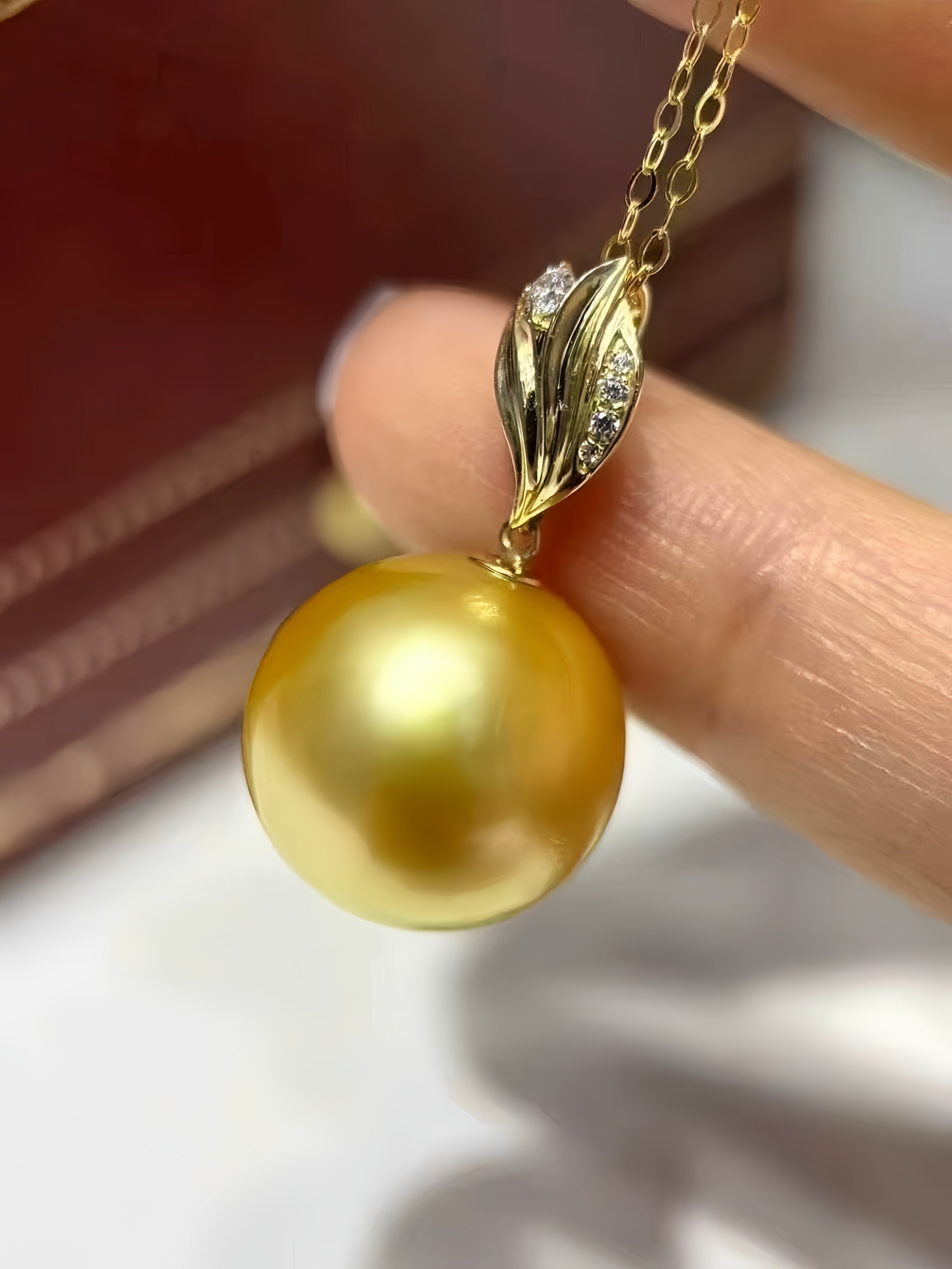 A close-up of a gold pendant necklace featuring a large, lustrous golden pearl suspended from a gold leaf-shaped setting with small clear stones, held between fingers.