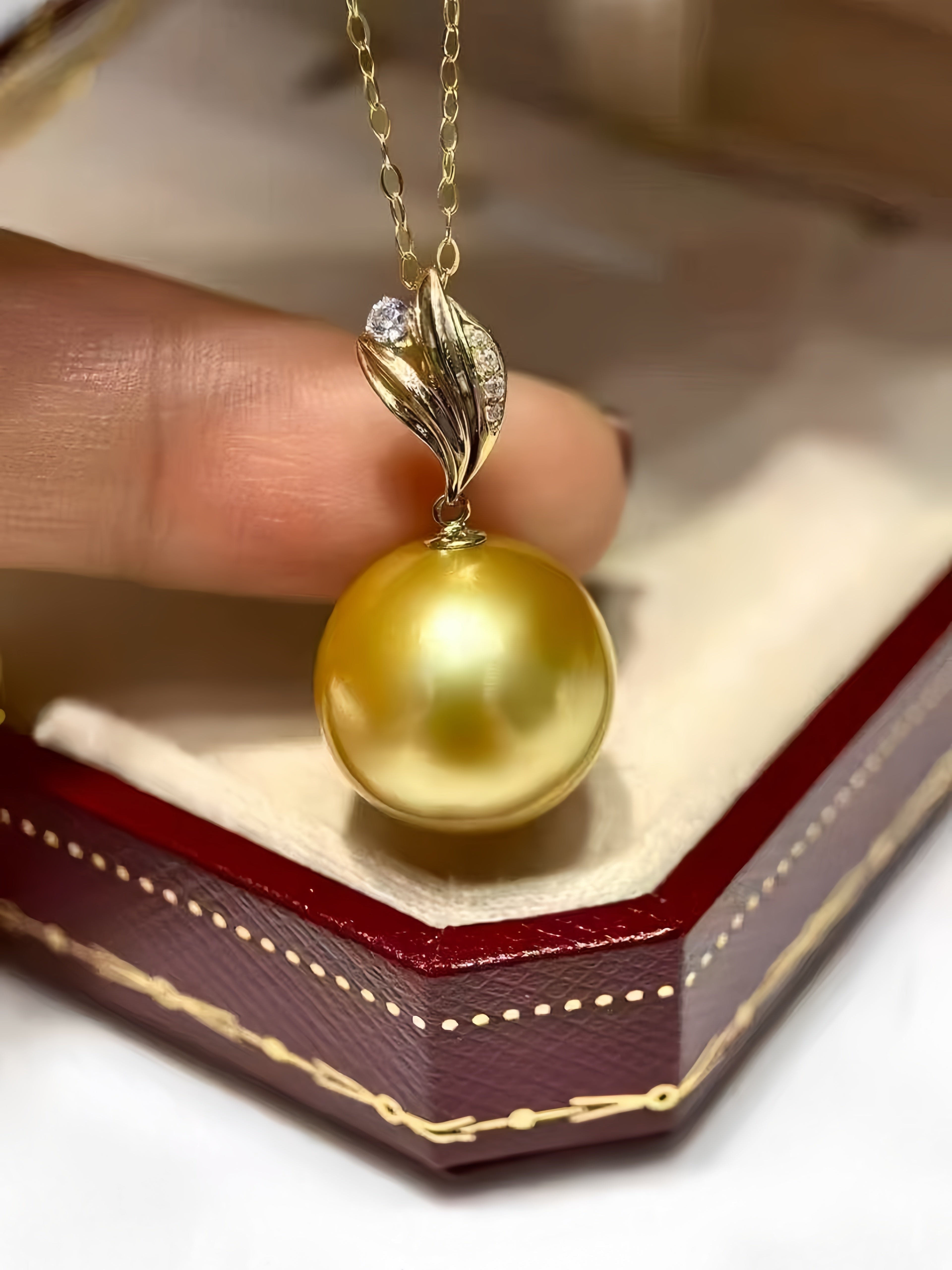 A gold pendant necklace featuring a large, lustrous yellow pearl pendant and a gold leaf-shaped setting with a small clear stone, held by a finger over an open red jewelry box.