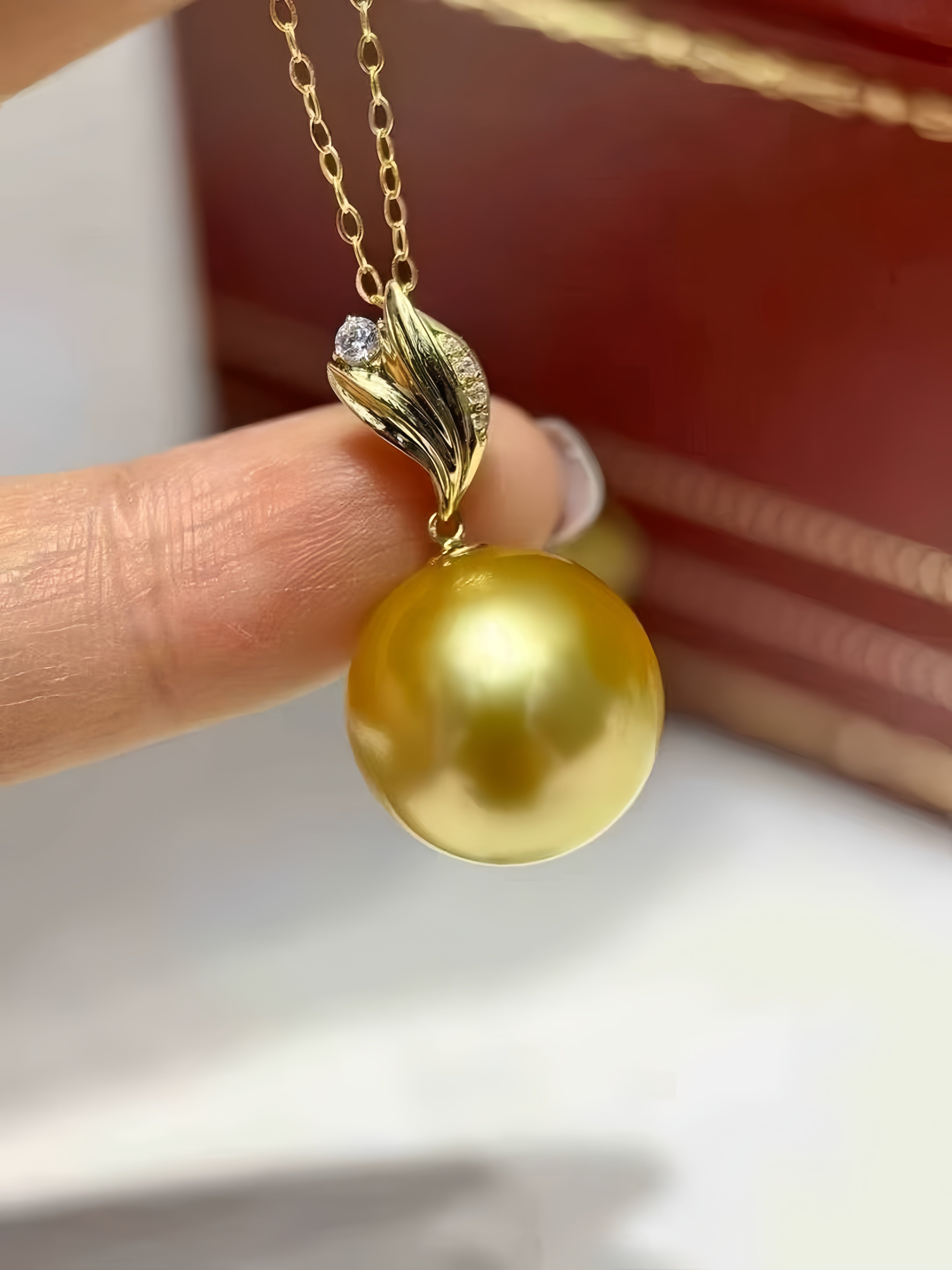 A close-up of a gold pendant necklace featuring a large, lustrous yellow pearl pendant and a gold leaf-shaped bail with a small clear stone, held by a person's fingers.