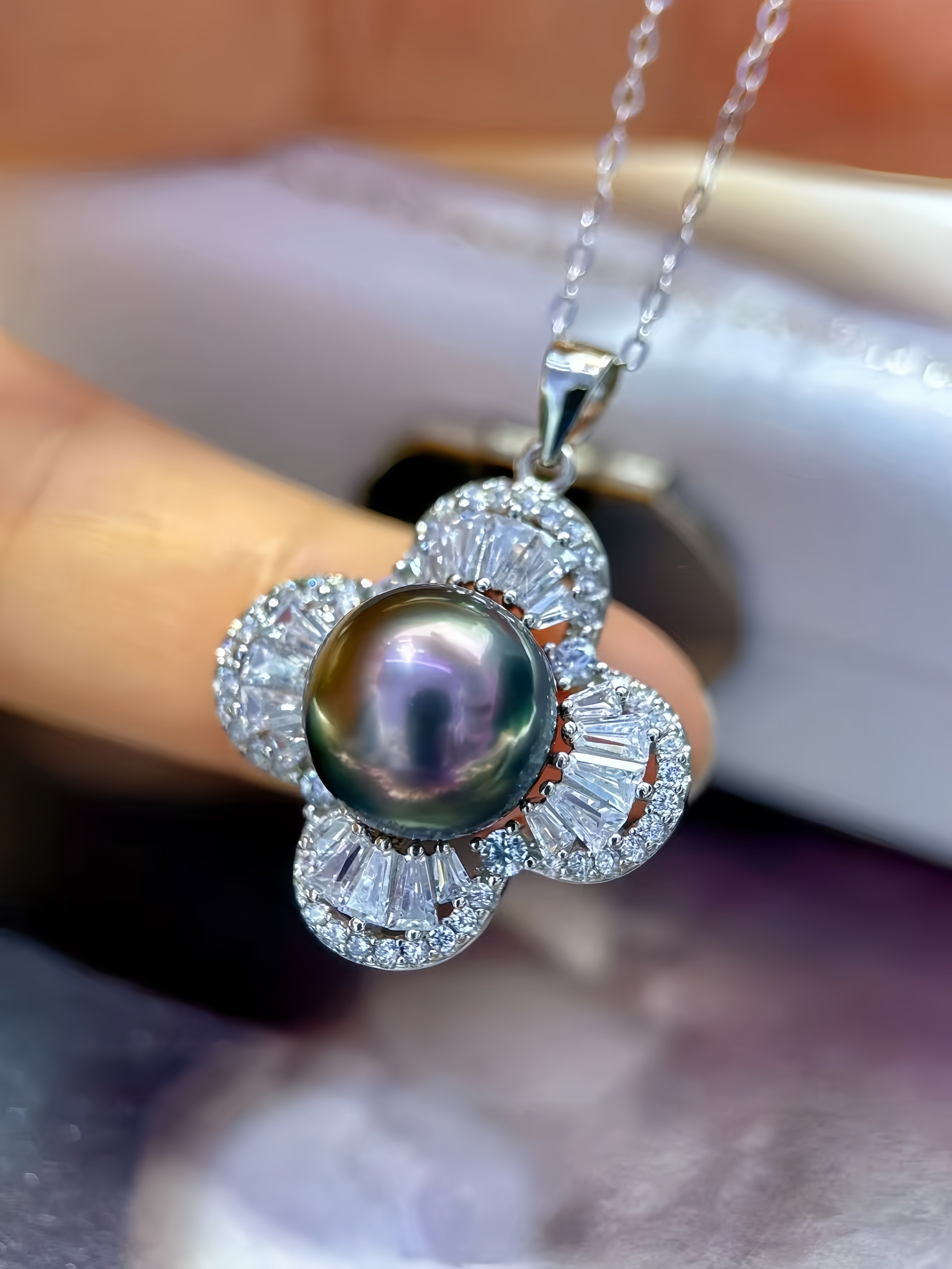 A close-up of a flower-shaped pendant necklace with a dark iridescent pearl in the center, surrounded by clear baguette and round stones in a silver-toned setting, held against a blurred background.