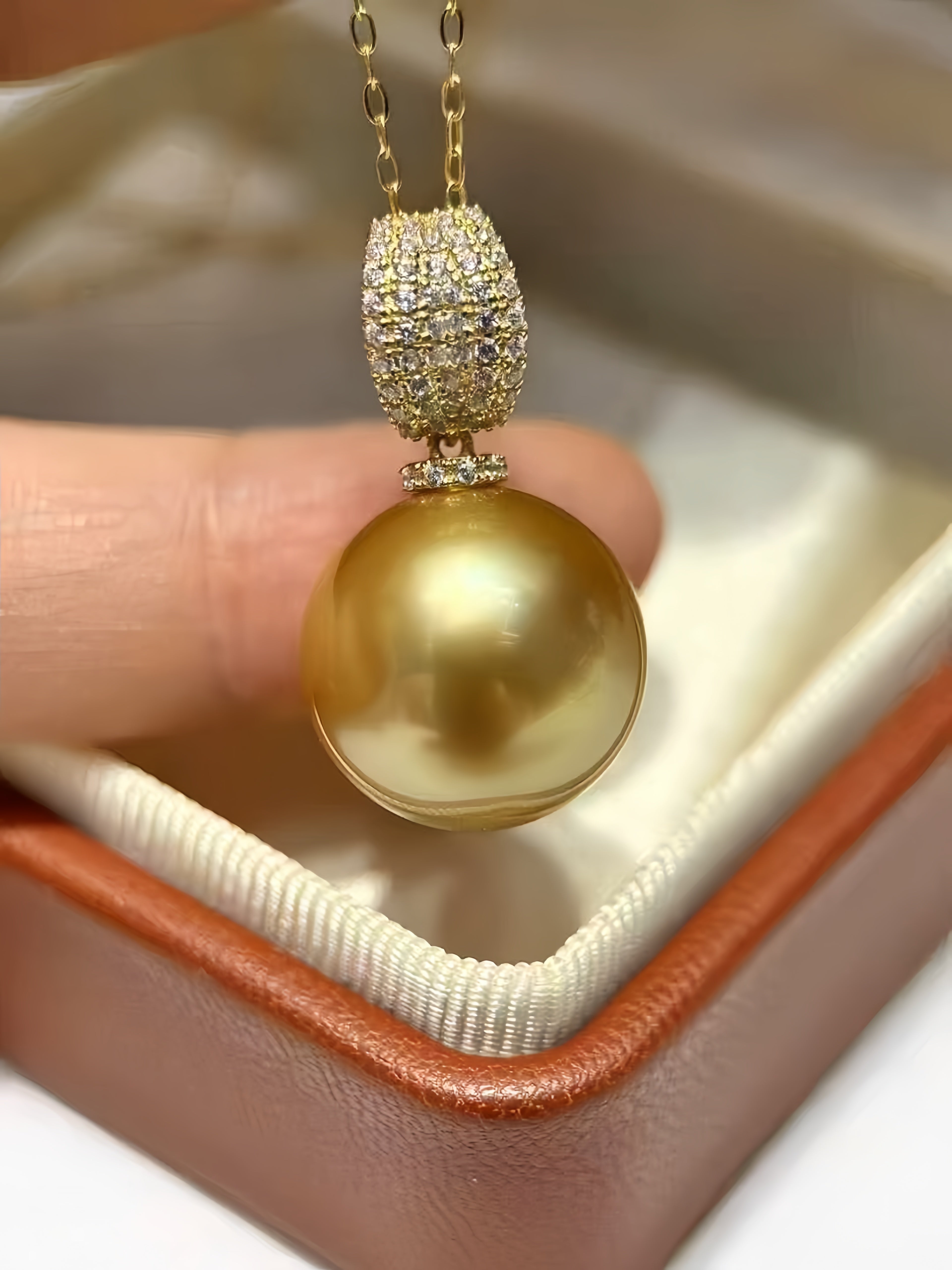 A close-up of a gold pendant necklace with a large round gold pearl and a diamond-studded spacer, held by a finger above an open brown jewelry box.