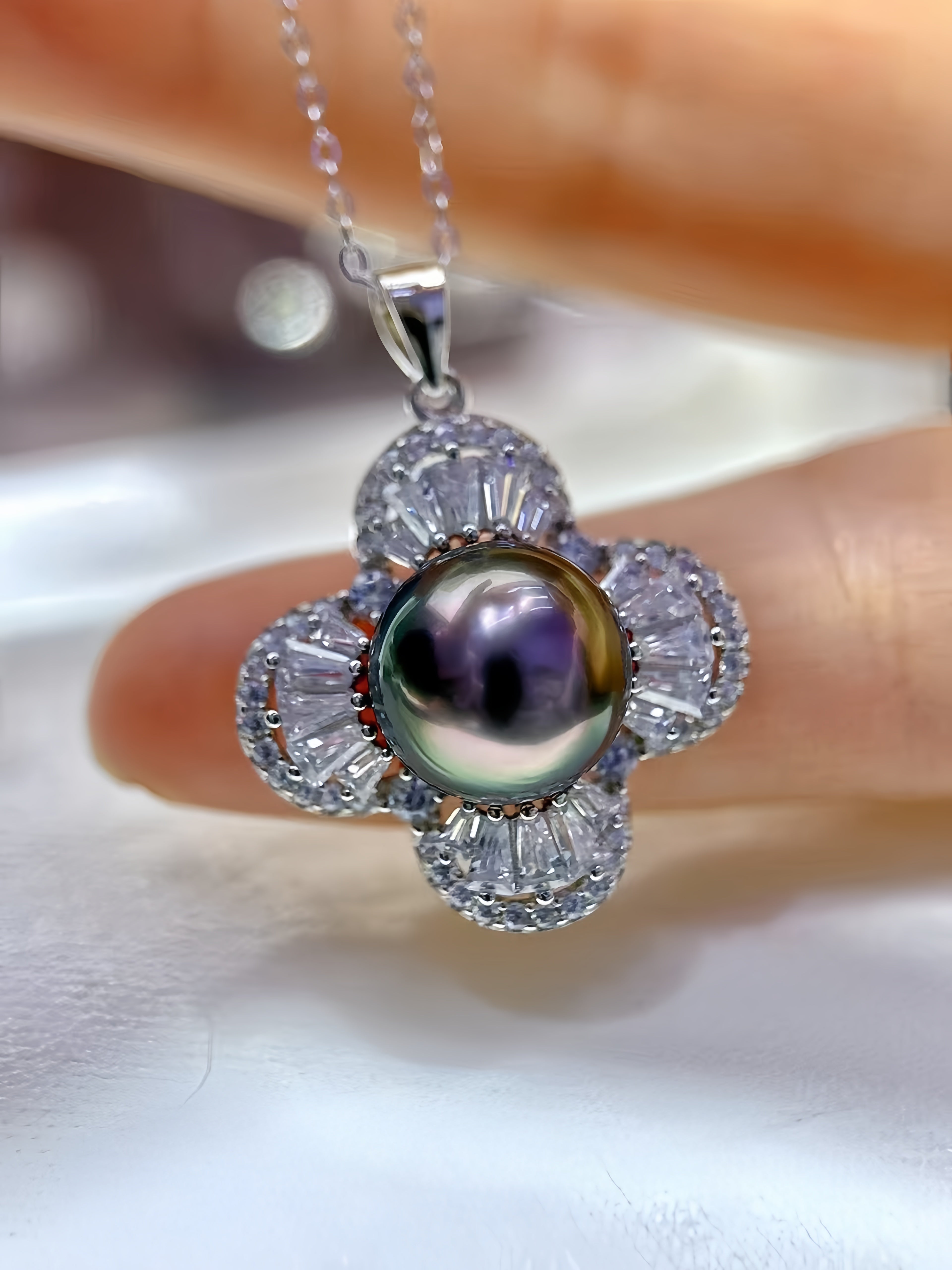 A close-up of a flower-shaped pendant necklace held between fingers. The pendant features a central iridescent freshwater pearl surrounded by clear baguette-cut stones in a silver-toned setting, with a matching silver chain visible at the top.