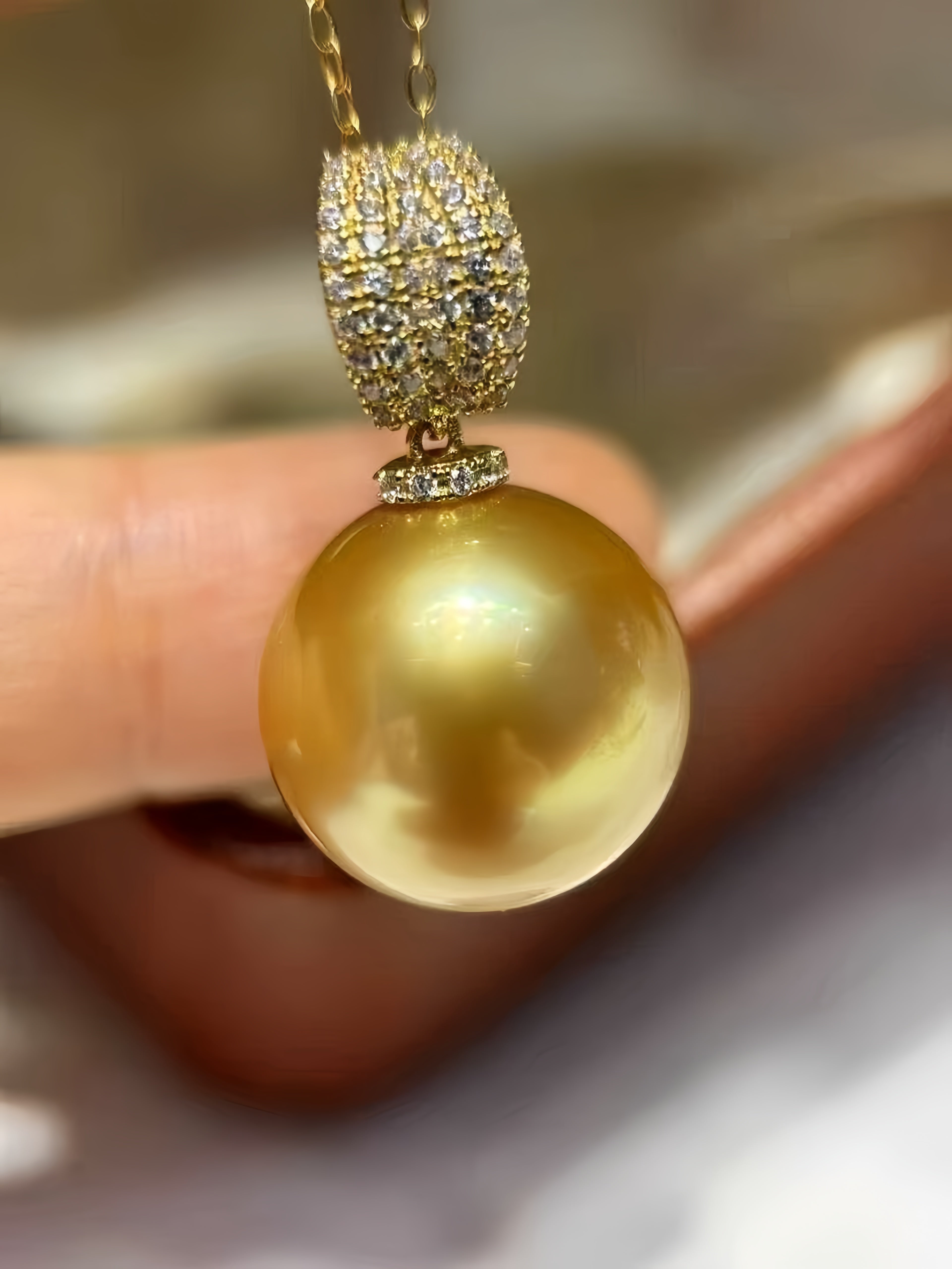 A close-up of a gold pendant necklace featuring a large, lustrous golden pearl and a pavé-set gold top, held by a person's fingers against a soft-focus background.