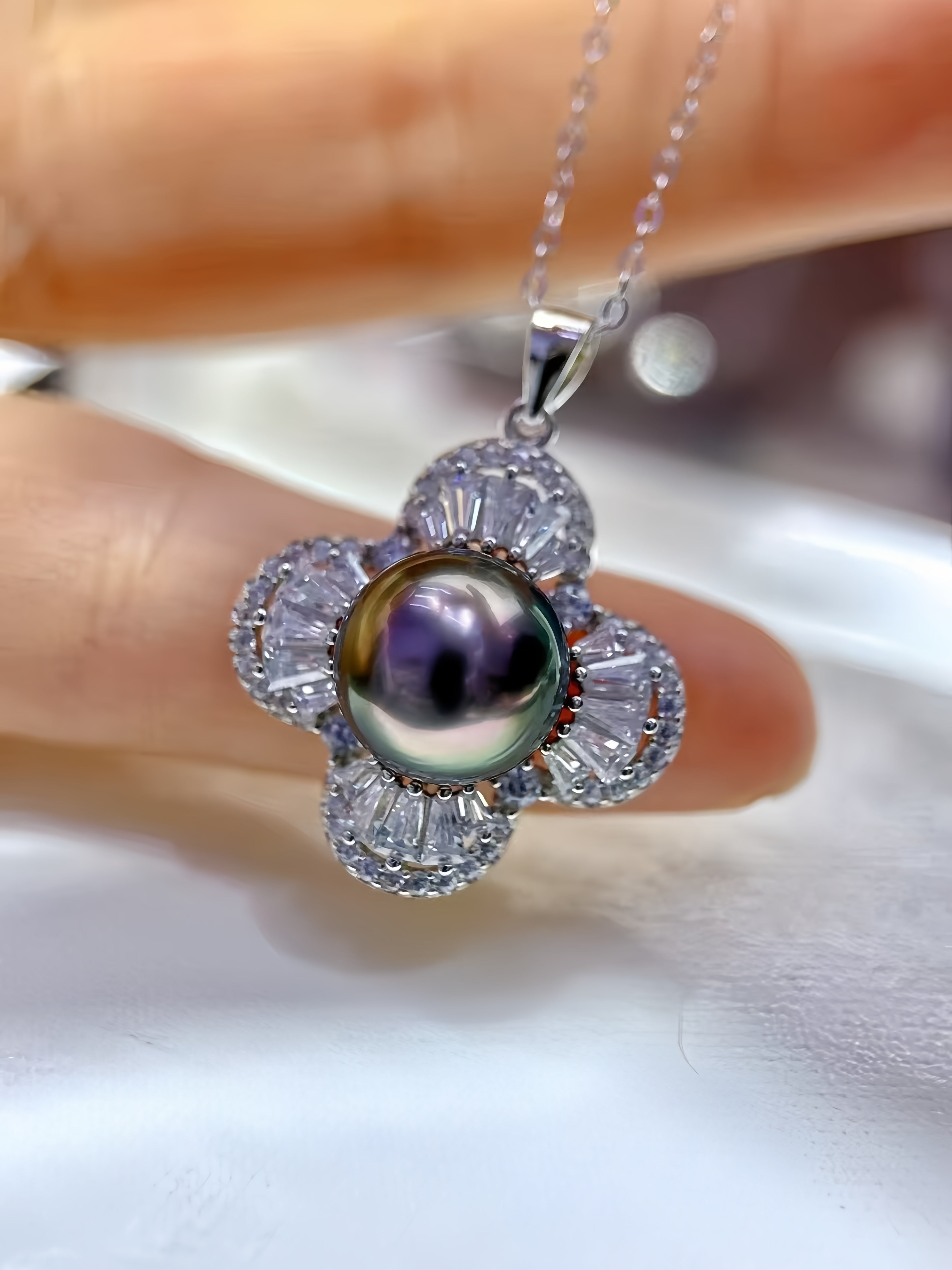 A close-up of a flower-shaped pendant necklace with a central iridescent pearl and clear baguette stones in a silver setting, held between fingers.