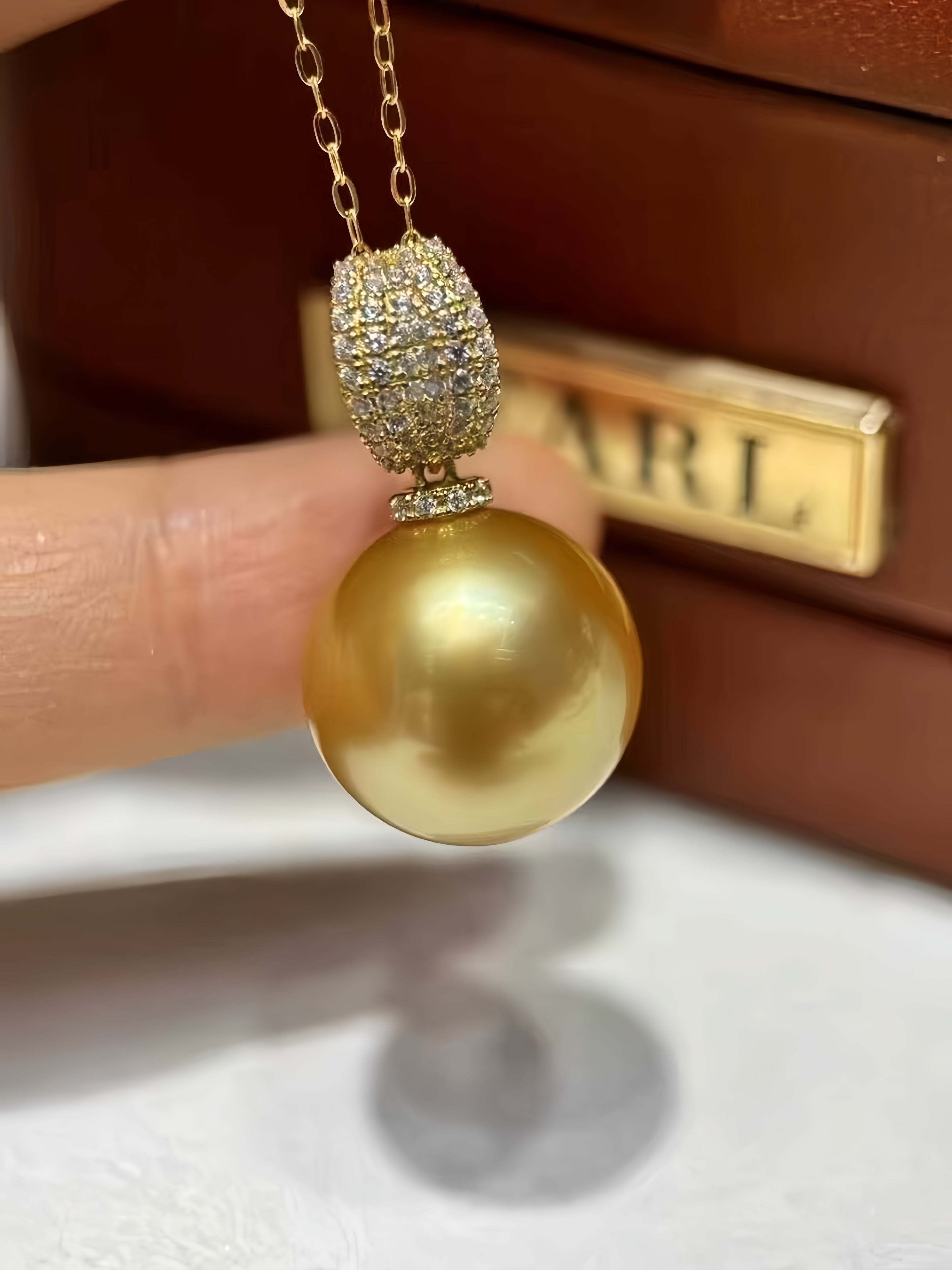 A gold pendant necklace featuring a large round gold pearl and a pavé-set top section, held by a hand in front of a brown jewelry box with a visible 'CARL' brand plaque.