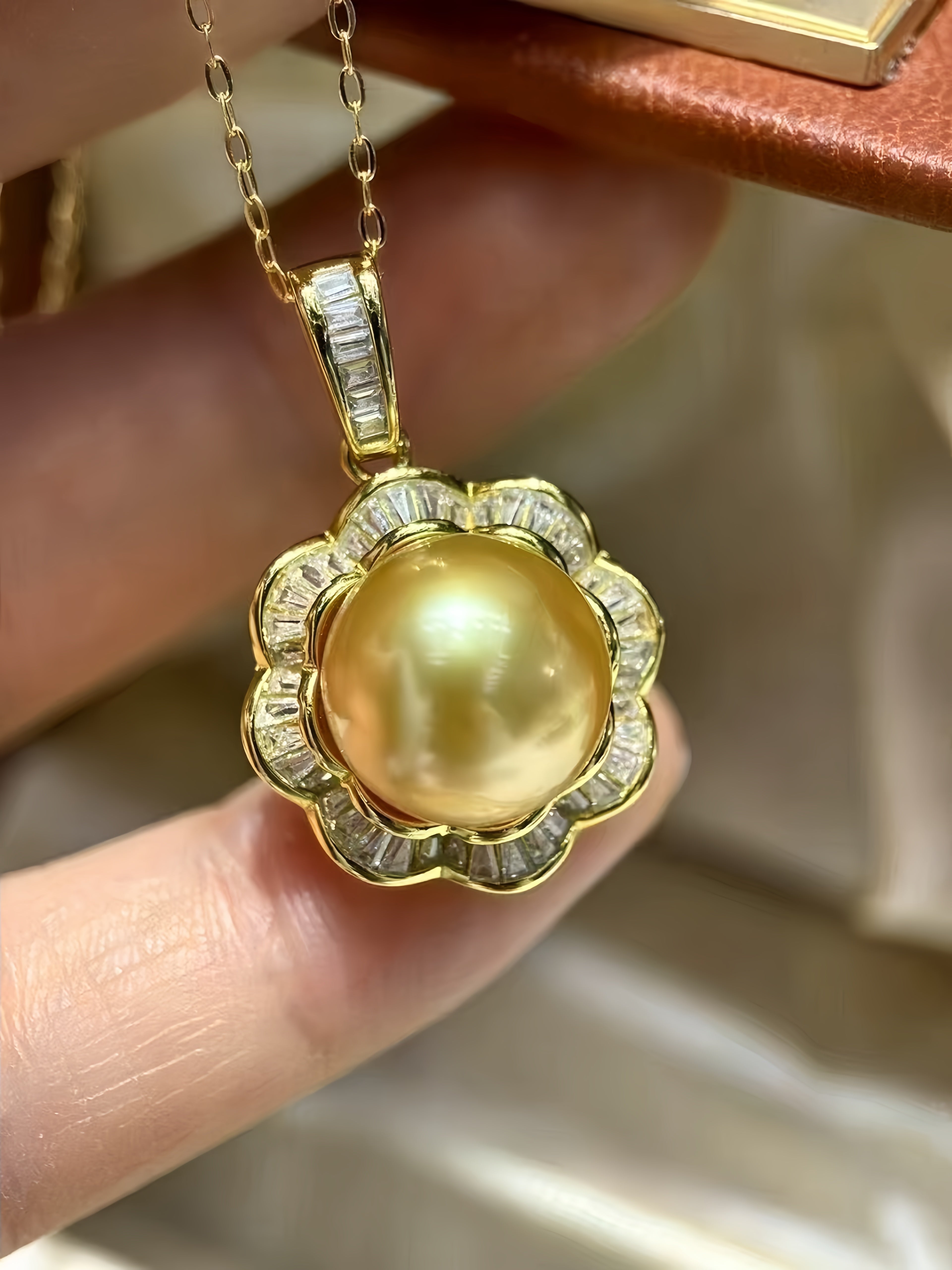 A close-up of a gold pendant necklace held by fingers. The pendant features a round, lustrous yellow-gold pearl surrounded by a scalloped border of small clear stones in a yellow gold setting, with a matching gold chain visible at the top.