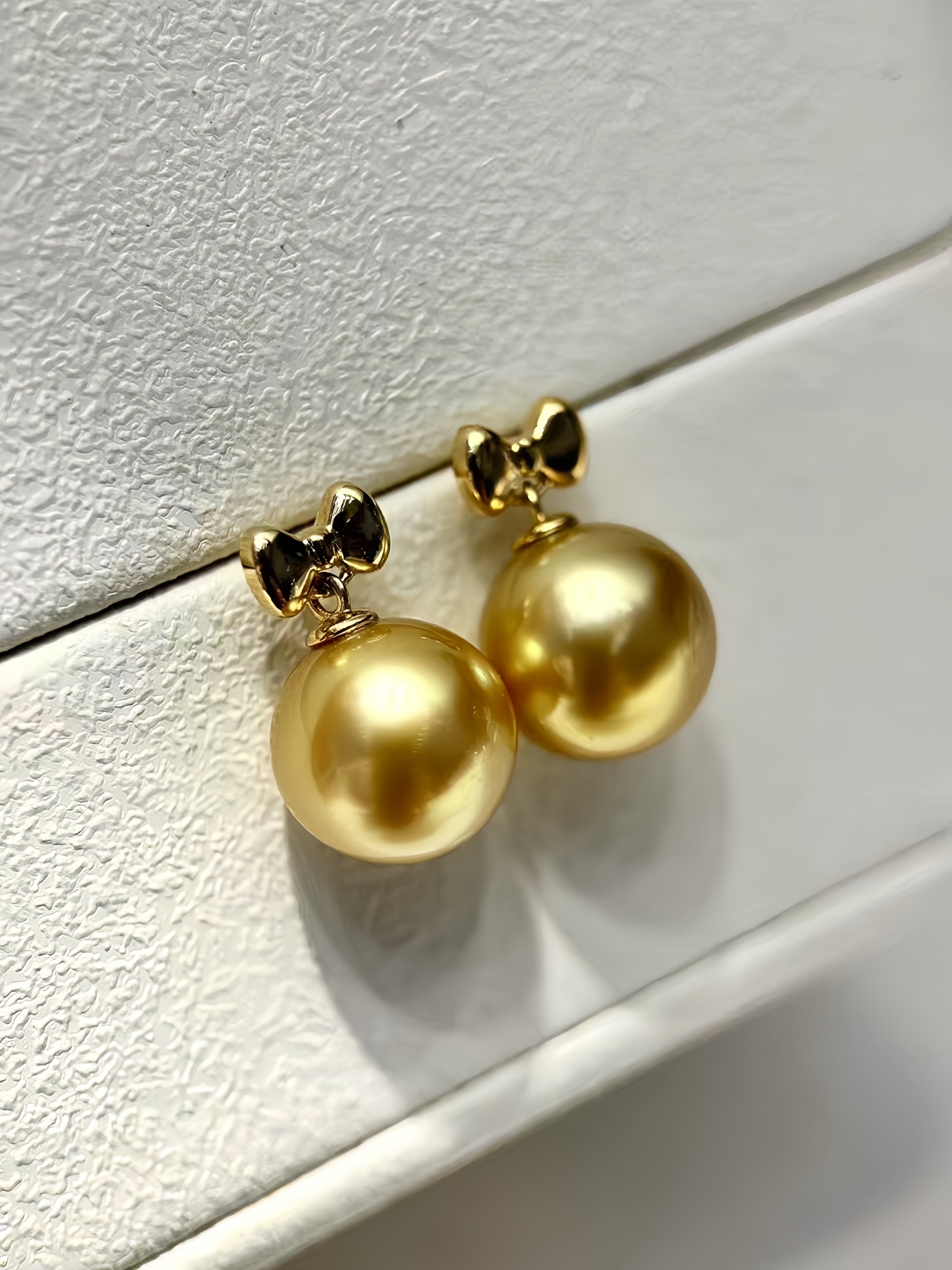 A pair of gold drop earrings with large round gold pearls and small bow-shaped gold settings, displayed on a textured white surface.