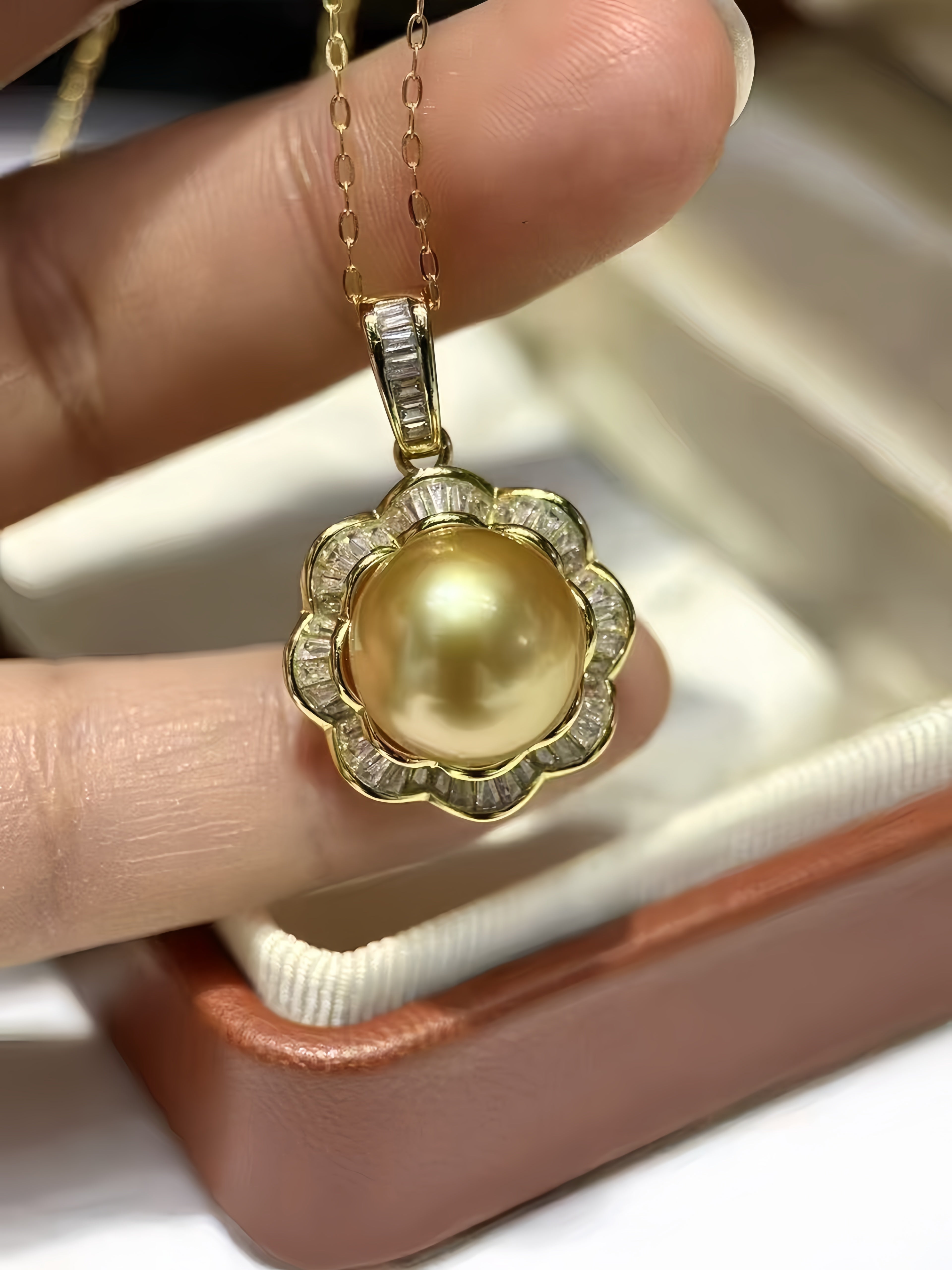 A close-up of a hand holding a gold pendant necklace with a large round pearl surrounded by small clear stones, with a jewelry box in the background.
