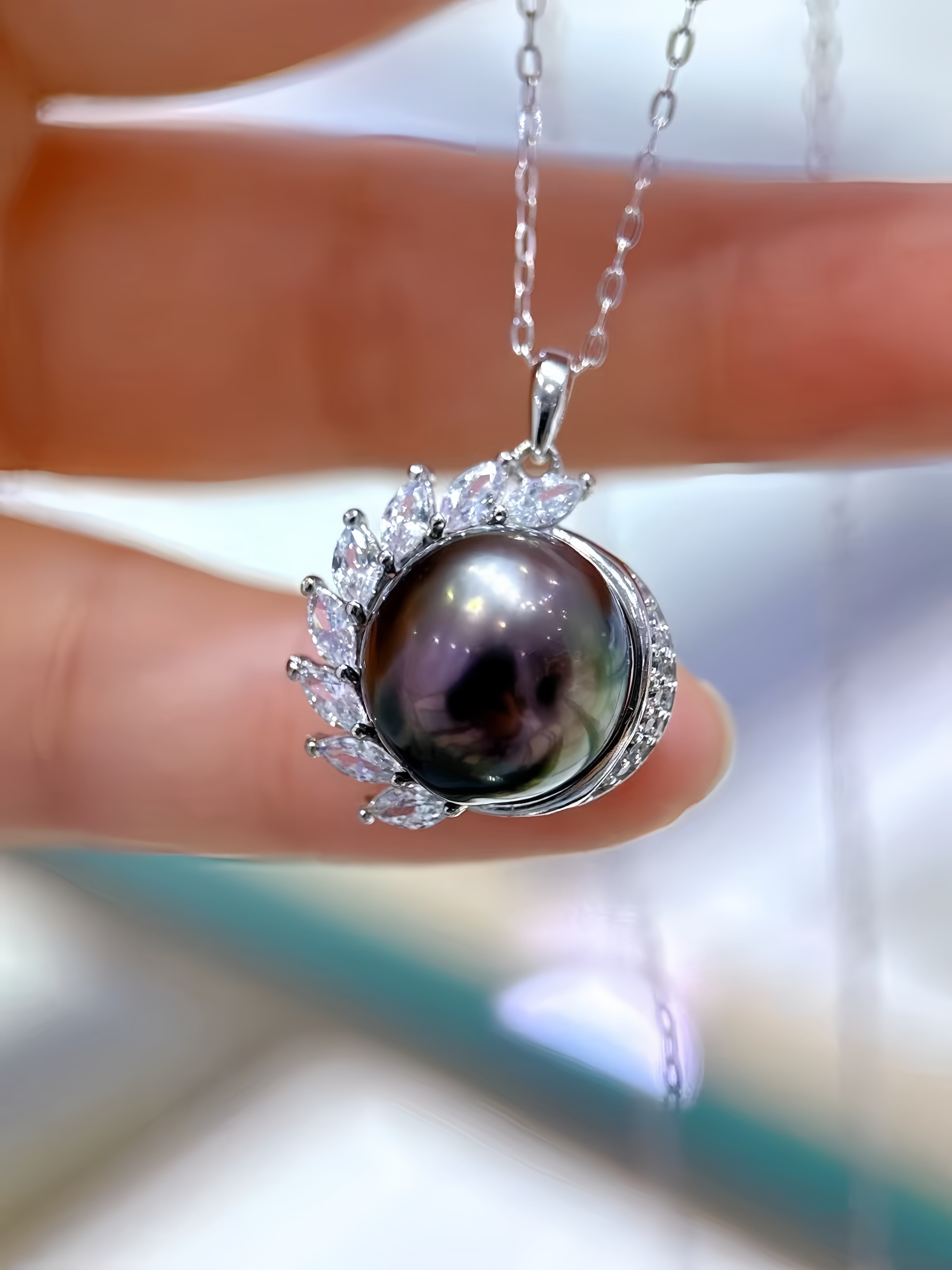 A close-up of a silver pendant necklace held between fingers. The pendant features a round iridescent dark pearl surrounded by small clear stones in a leaf-like halo, with a silver chain extending upward.