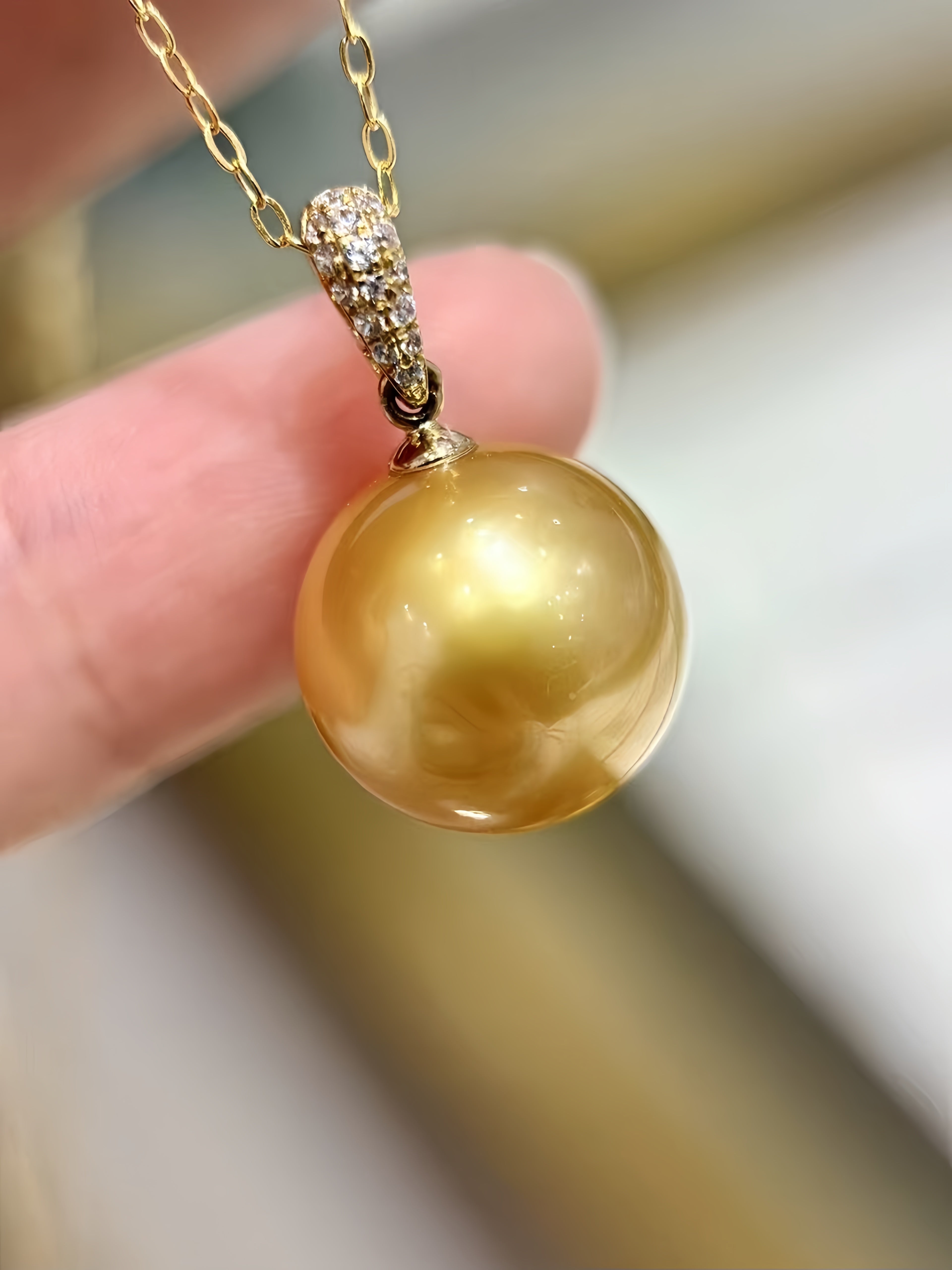 A close-up of a gold pendant necklace featuring a large, lustrous golden pearl pendant and a pavé-set gold bail, held by a hand against a softly blurred background.