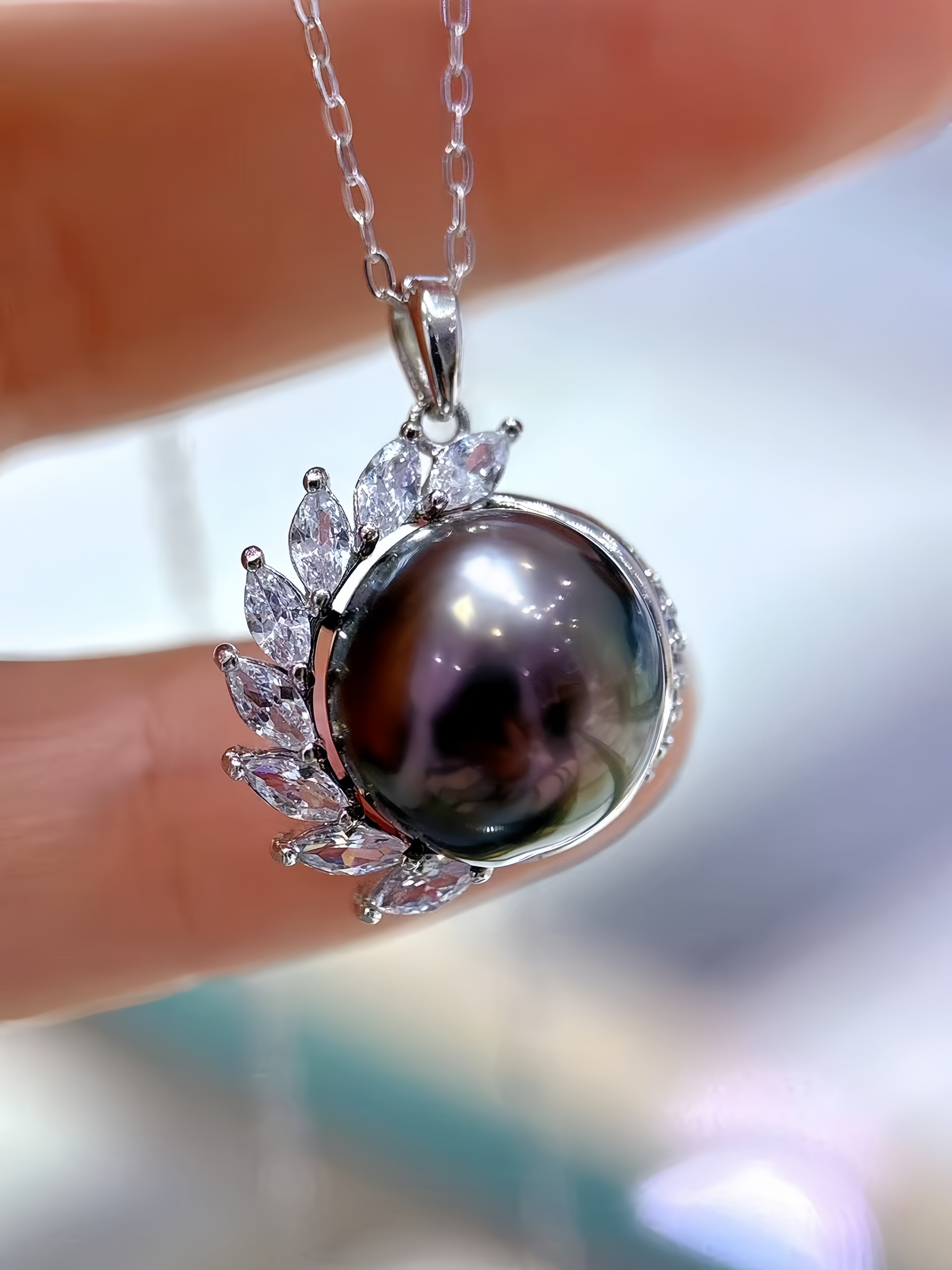 A close-up of a silver pendant necklace featuring a large, glossy dark pearl surrounded by clear marquise-cut crystals, held against a blurred background.
