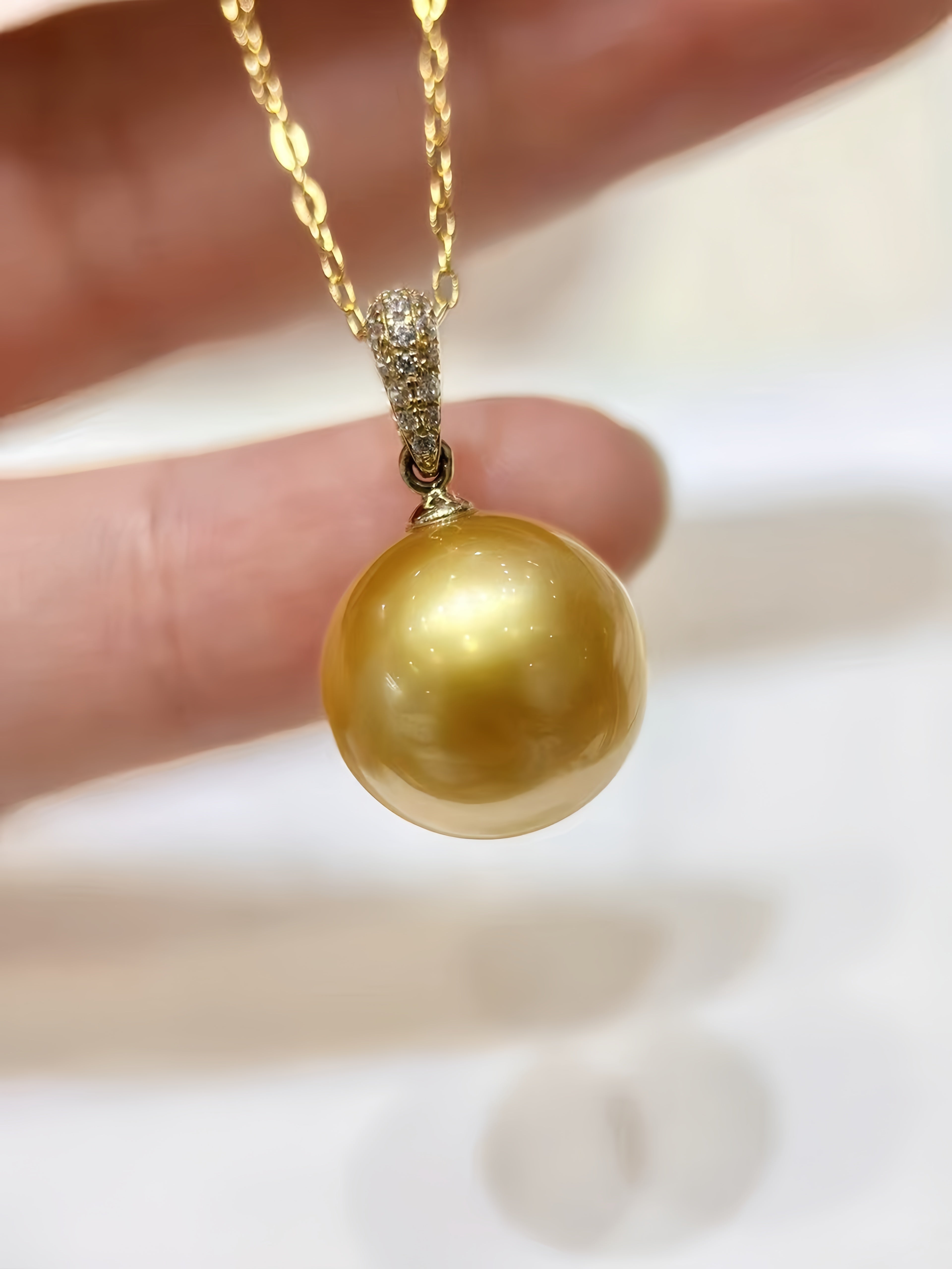 A gold pendant necklace featuring a large, round, lustrous gold freshwater pearl, held by a hand against a bright white background.
