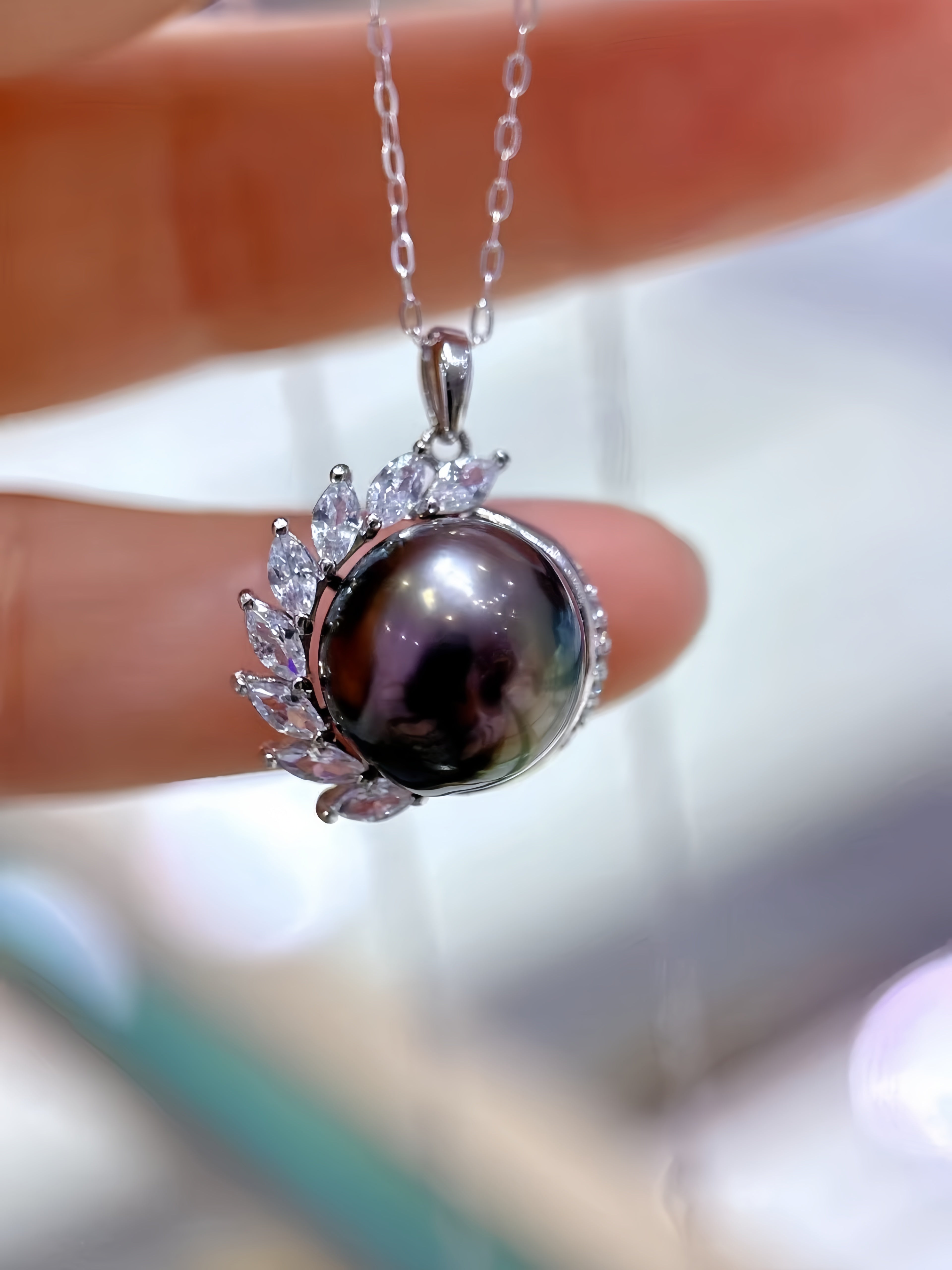 A close-up of a silver pendant necklace held by a hand. The pendant features a large, round, dark pearl with a pearlescent sheen, surrounded by a silver leaf-like halo accented with small clear stones.