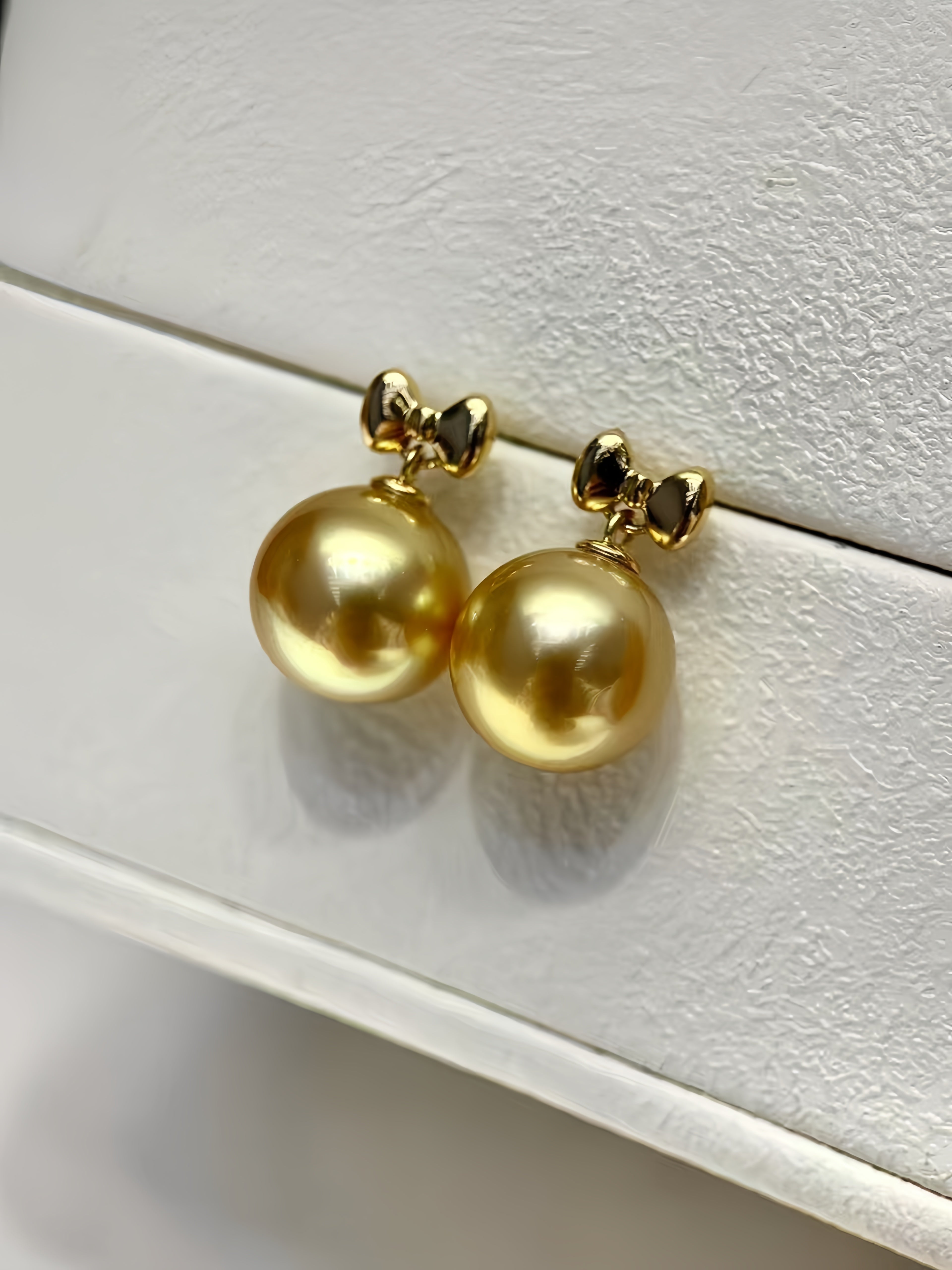 A pair of gold stud earrings with large round gold freshwater pearls and small bow-shaped post details, displayed on a textured white jewelry box.