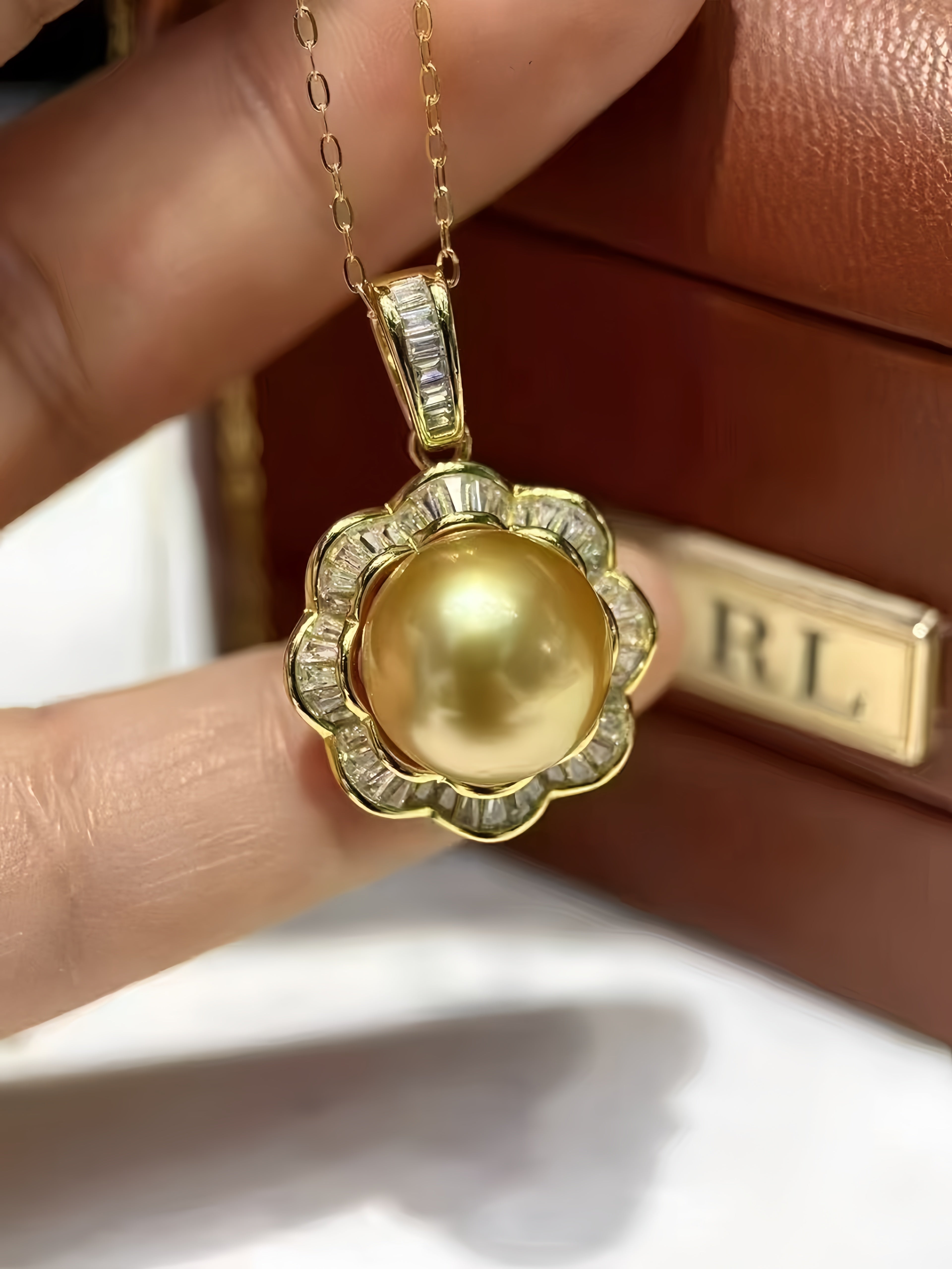 A gold pendant necklace with a large round pearl surrounded by small clear stones, held between fingers in front of a brown leather box with an RL logo.