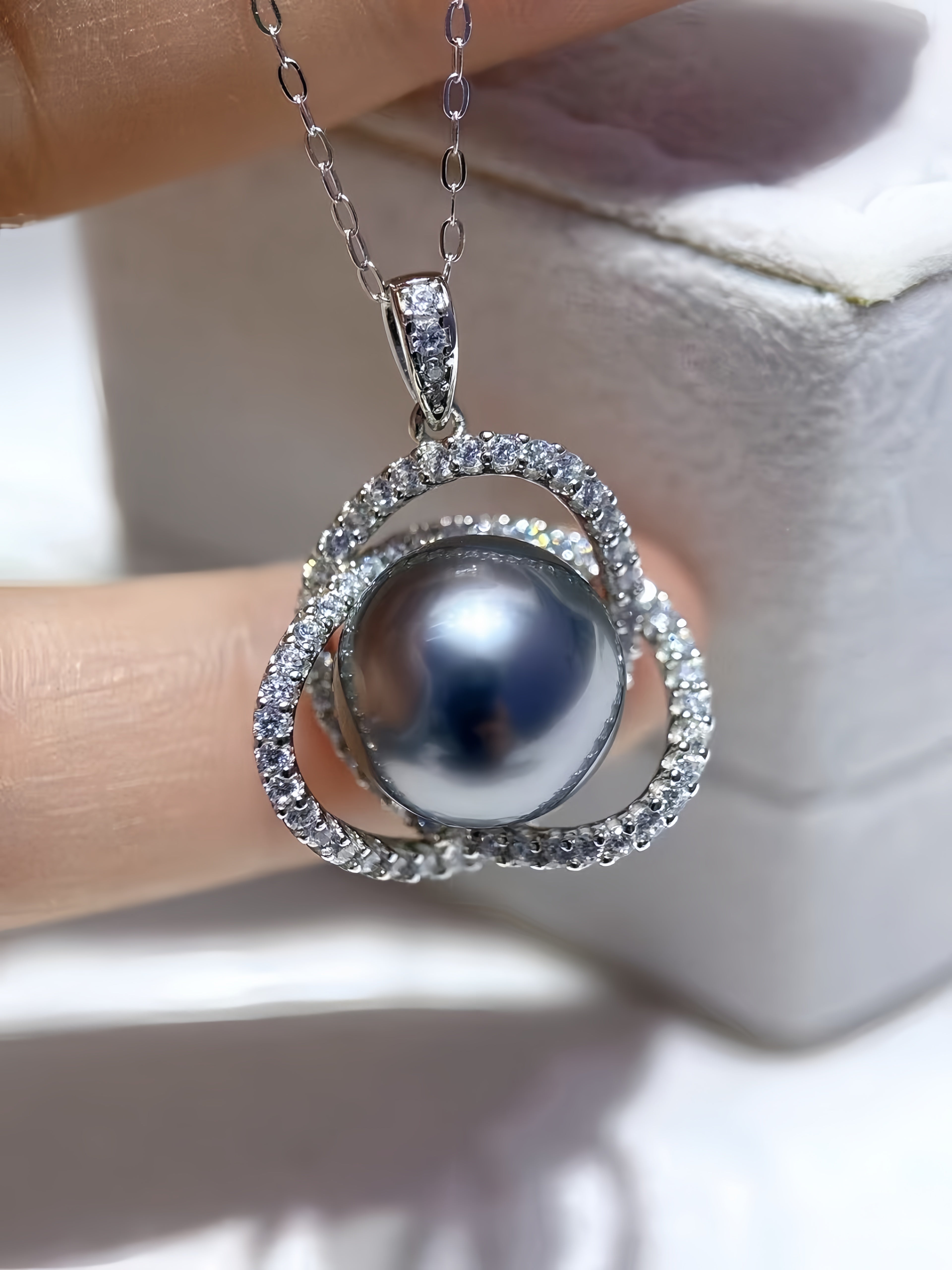A silver pendant necklace featuring a large dark gray pearl surrounded by a double halo of small clear pavé stones, held by a hand in front of a white jewelry box.