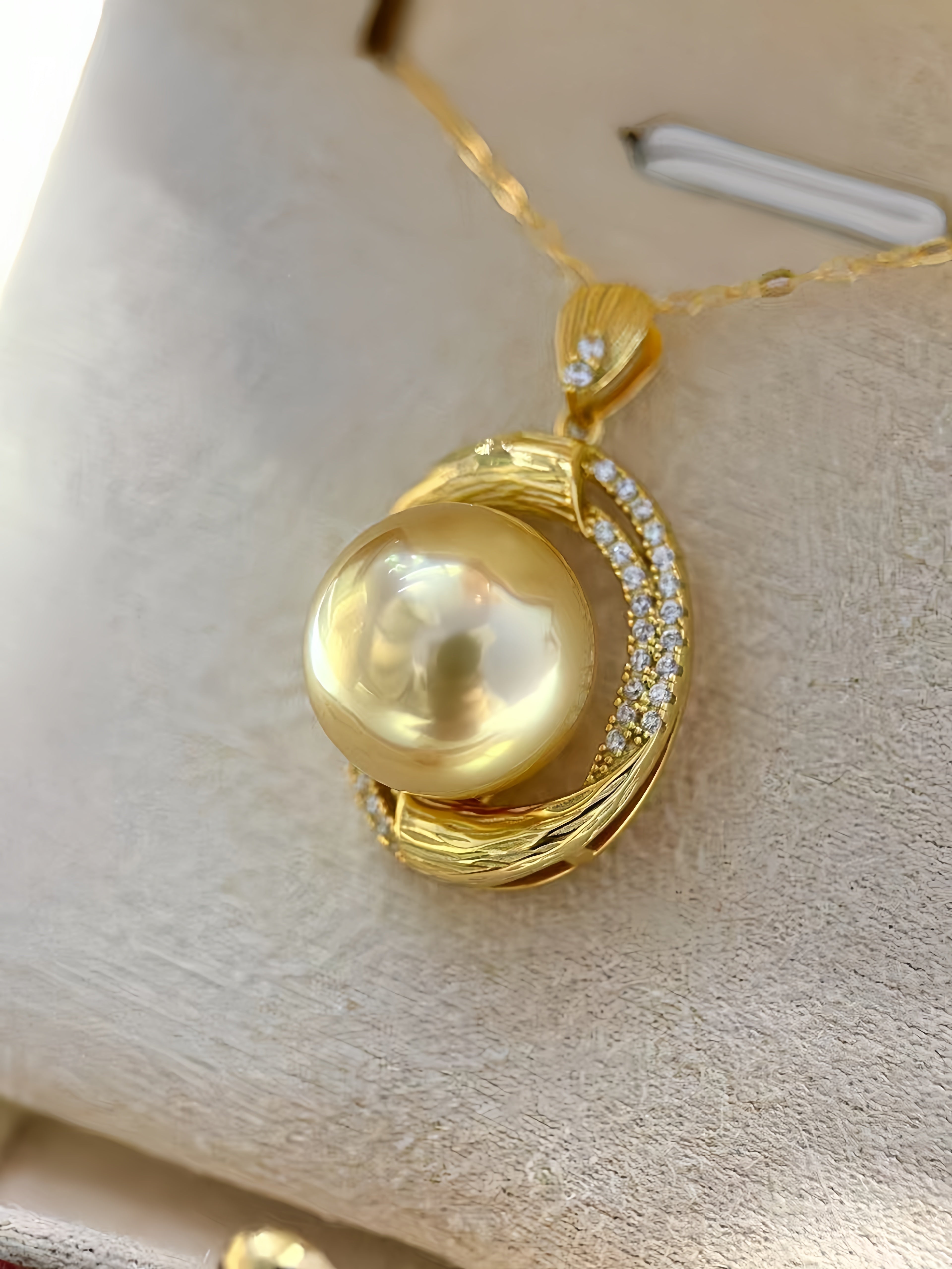 A gold pendant necklace featuring a large, lustrous freshwater pearl surrounded by a crescent-shaped setting accented with small clear stones, displayed on a beige jewelry stand.