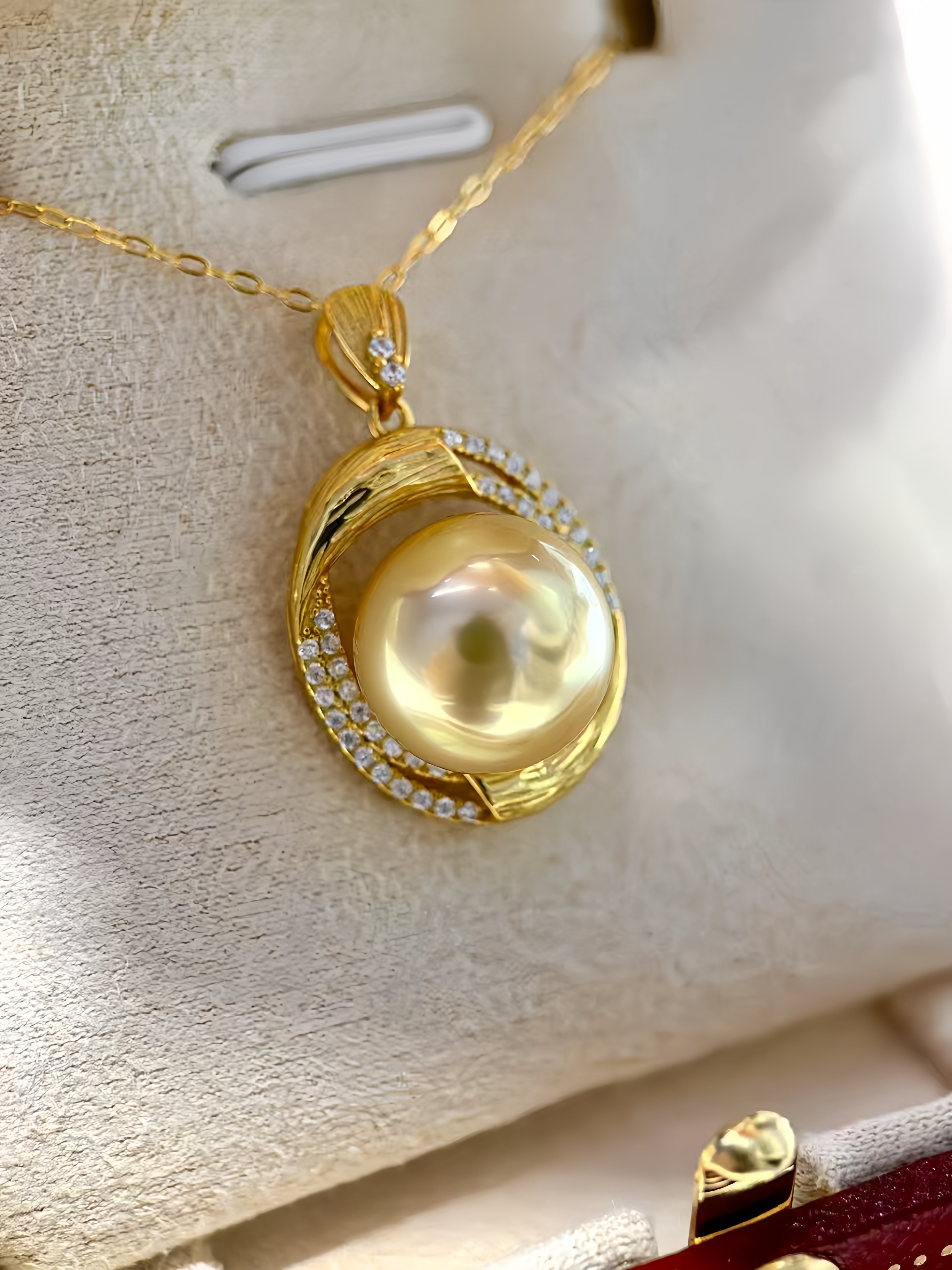 A gold pendant necklace featuring a large, lustrous pearl surrounded by a halo of small clear stones, displayed on a cream-colored jewelry box.