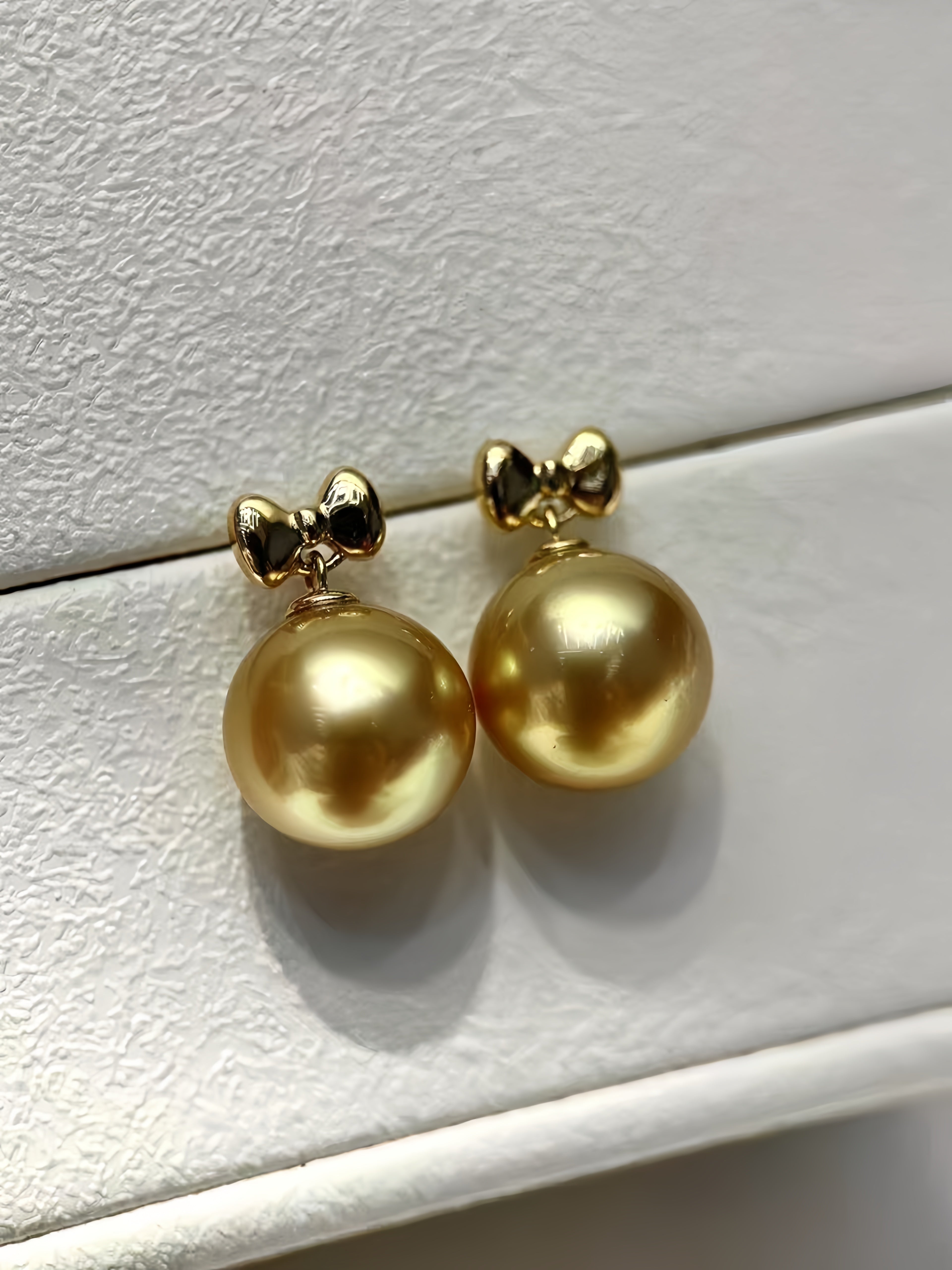 A pair of gold-tone stud earrings with a small bow-shaped top and a large round gold pearl drop, displayed on a white textured surface.