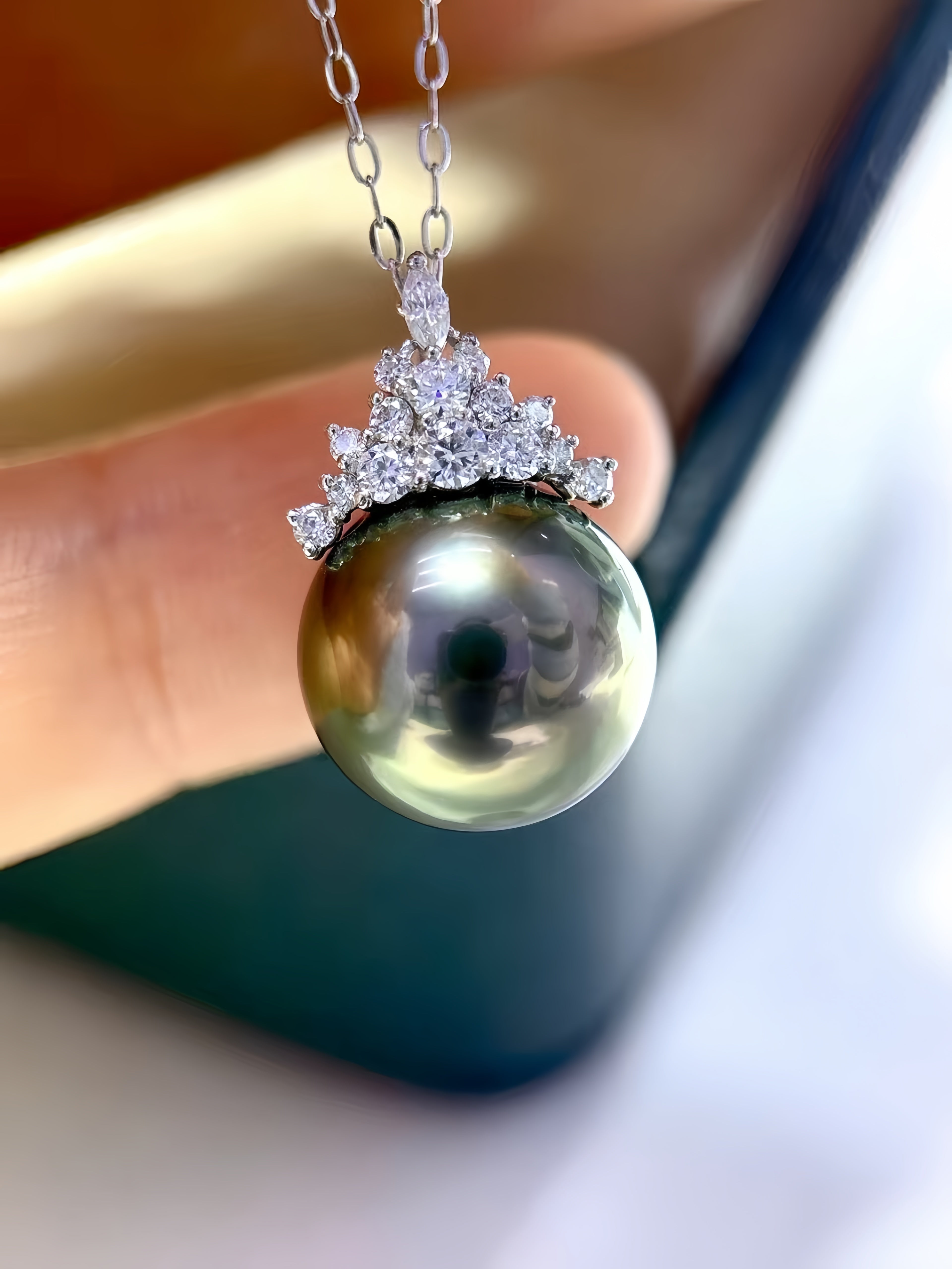 A close-up of a freshwater pearl pendant necklace held by a hand. The pendant features a large, round olive-green pearl with a silver-colored floral crown set with small clear stones, suspended from a silver link chain.