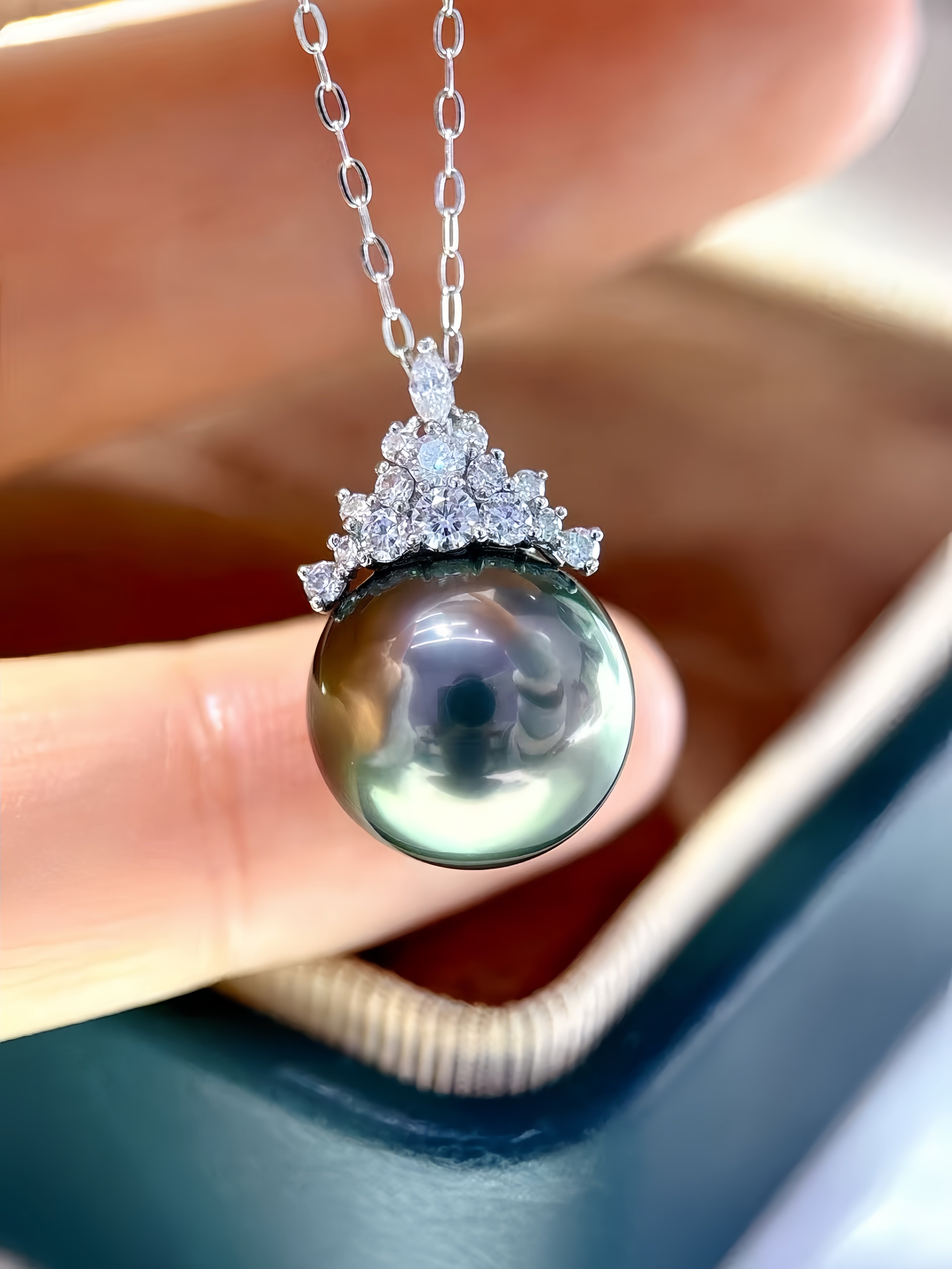 A close-up of a silver pendant necklace featuring a large, round, dark green-black pearl with a halo of small clear stones, held by fingers above an open jewelry box.
